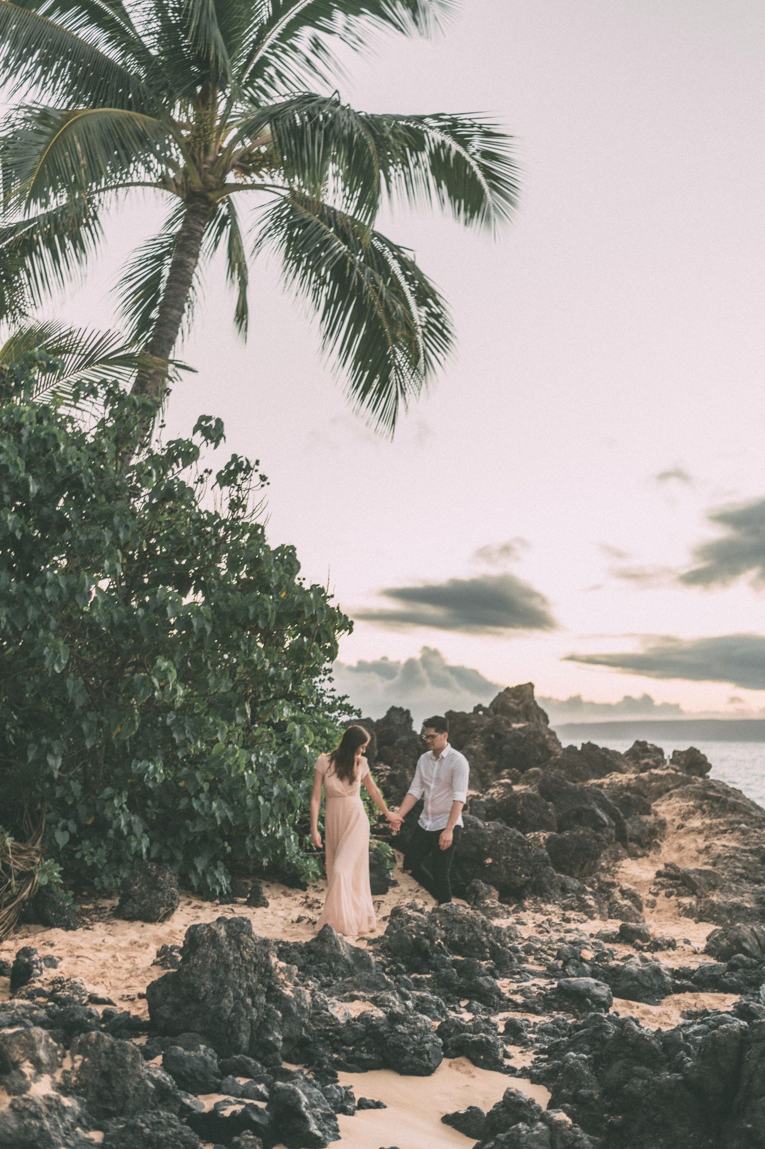 Couple holding hands on rocky beach with palm trees and sunset sky behind them.