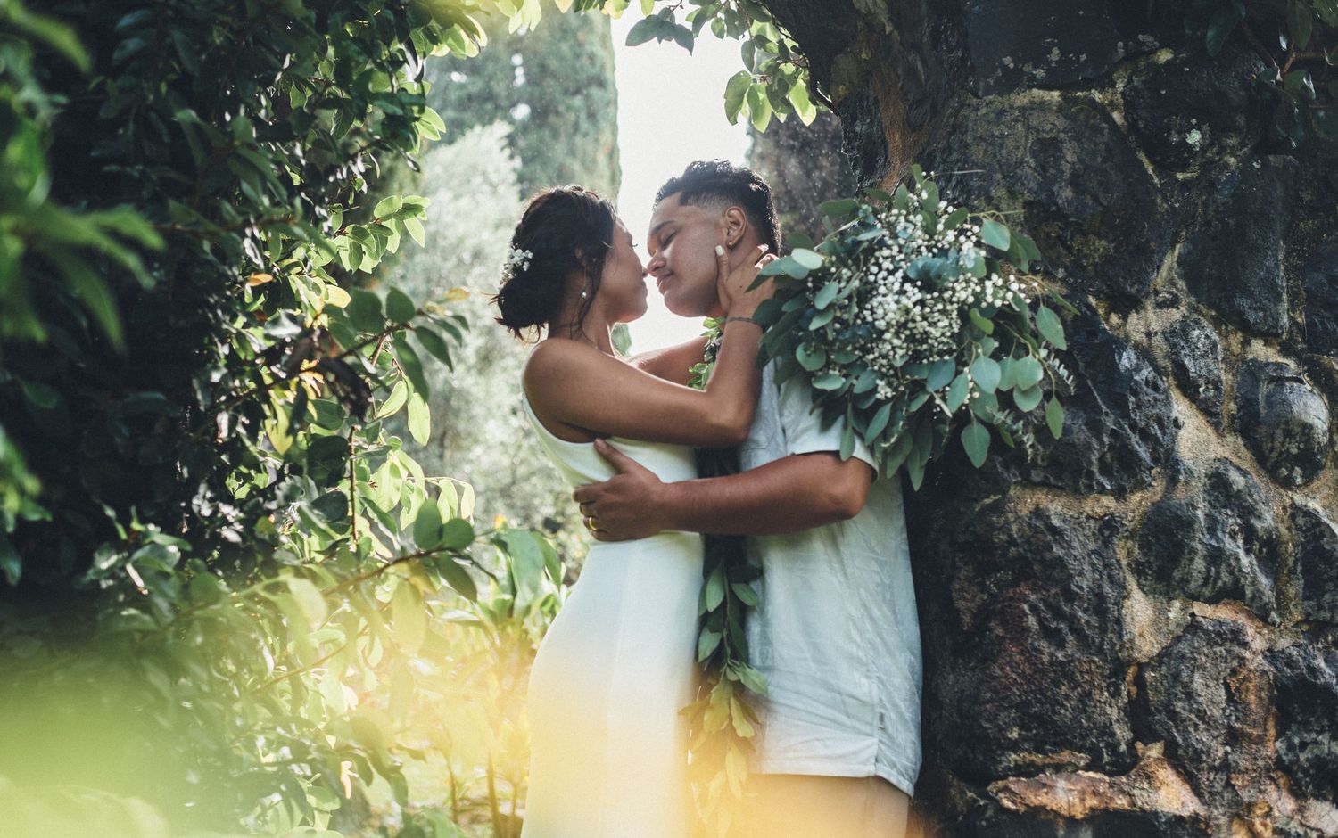 Two brides share a romantic kiss surrounded by lush greenery and stone walls, one holding a white floral bouquet.