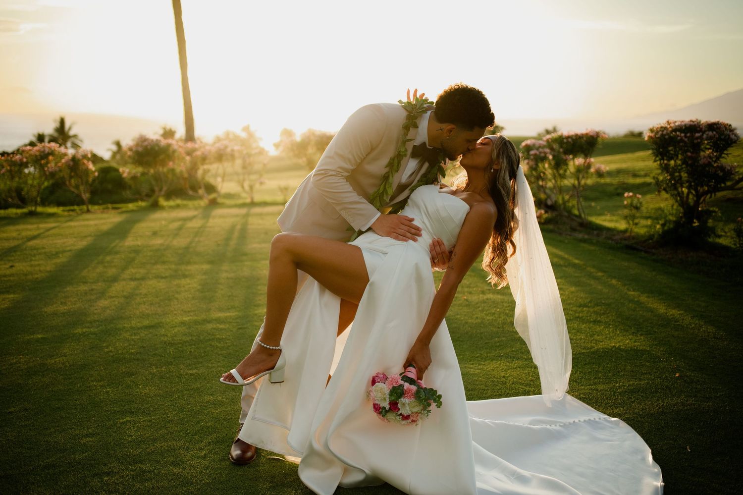 Bride and groom share a romantic dip kiss at golden hour on a lush green lawn, with bride holding a pink bouquet.