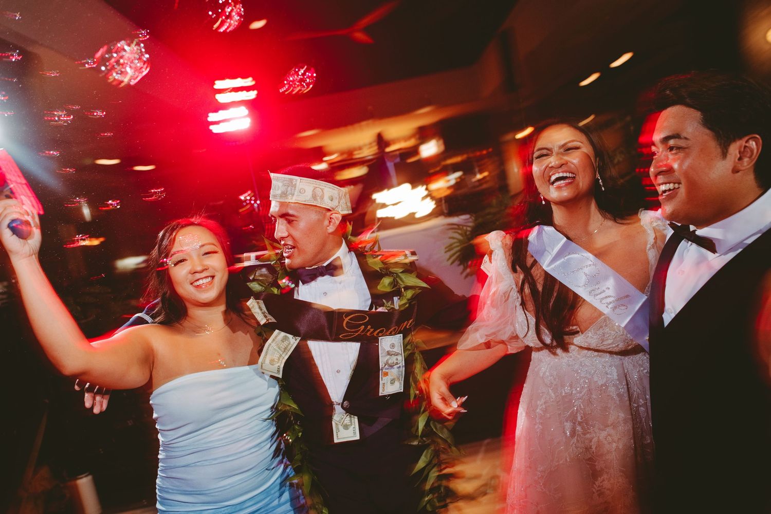 Joyful wedding reception guests dancing and celebrating under red lights, wearing leis and formal attire.