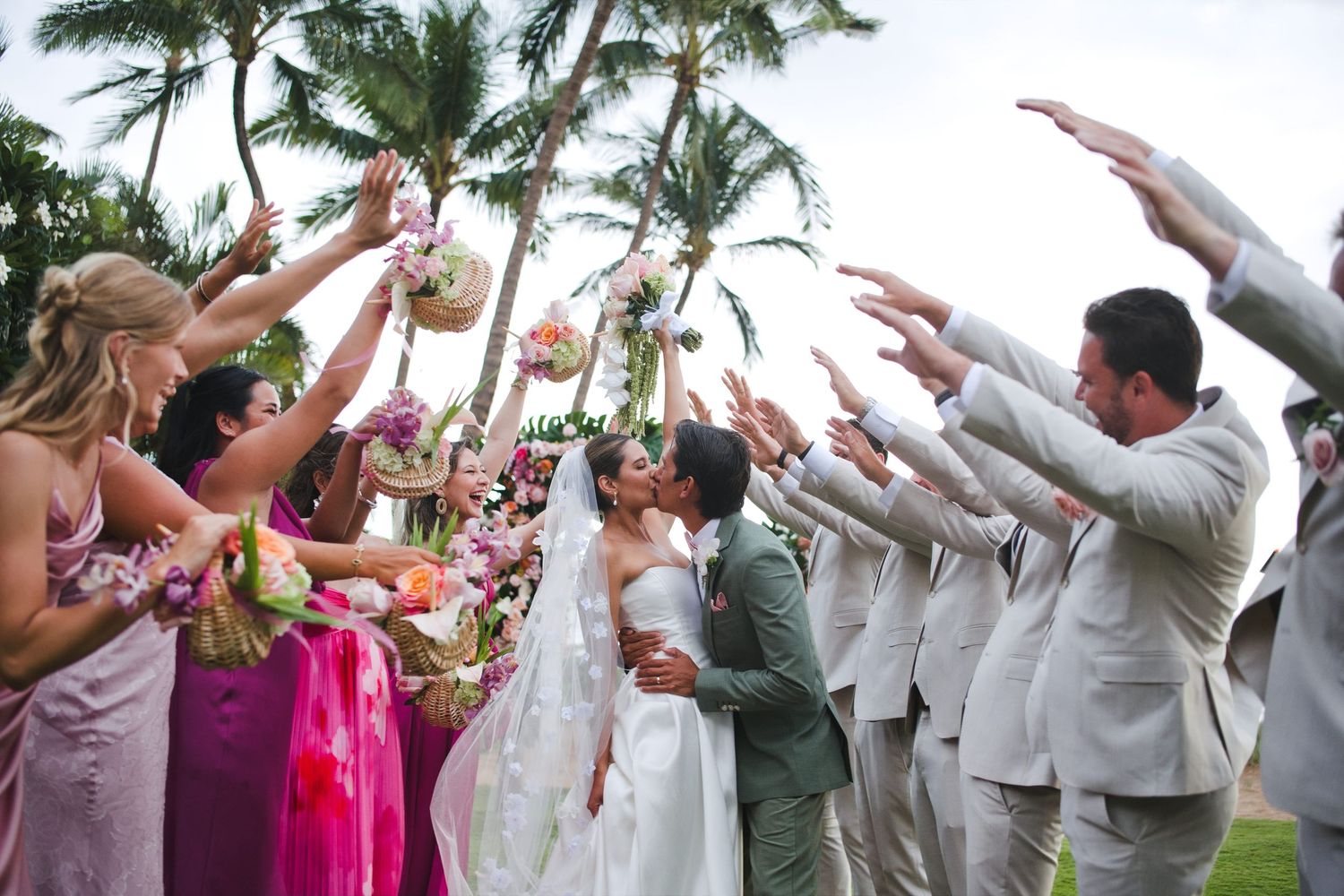 Bride and groom share a kiss under an arch of raised arms from wedding party guests at a tropical outdoor ceremony.