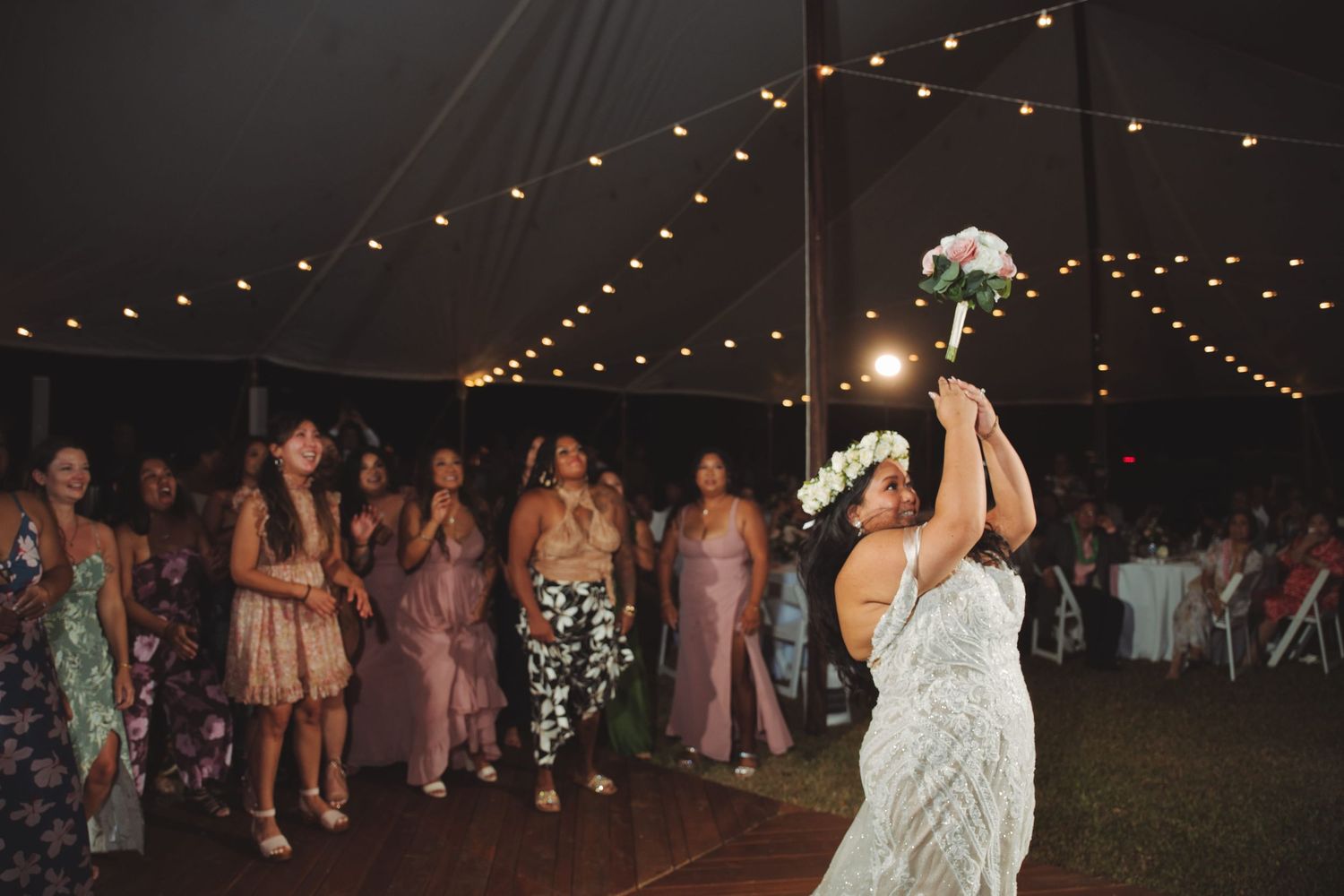 Bride in lace gown tossing bouquet to guests under string lights at outdoor wedding reception.
