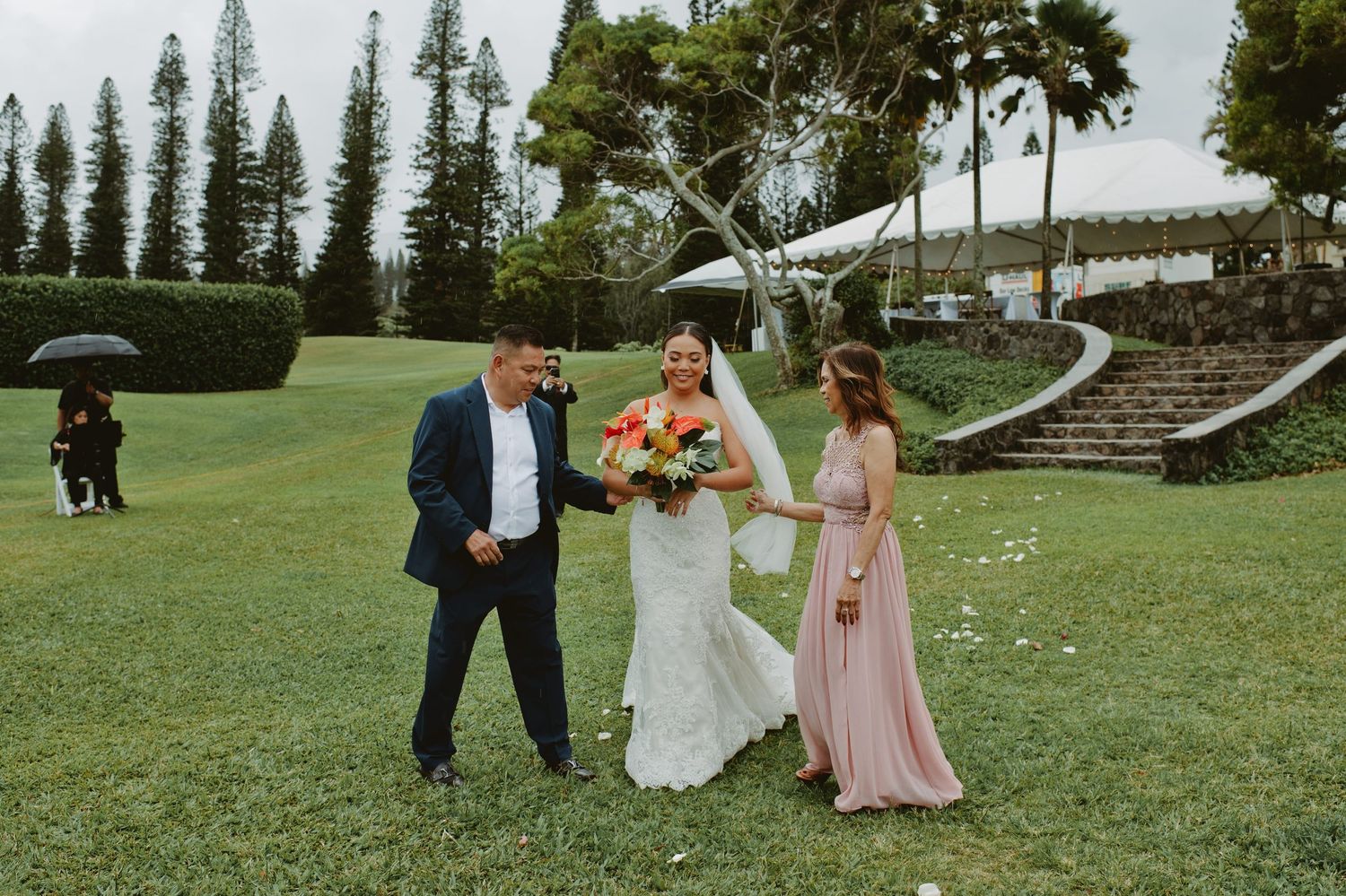 Bride in white gown walks with groom and bridesmaid in pink dress on green lawn at outdoor wedding venue.