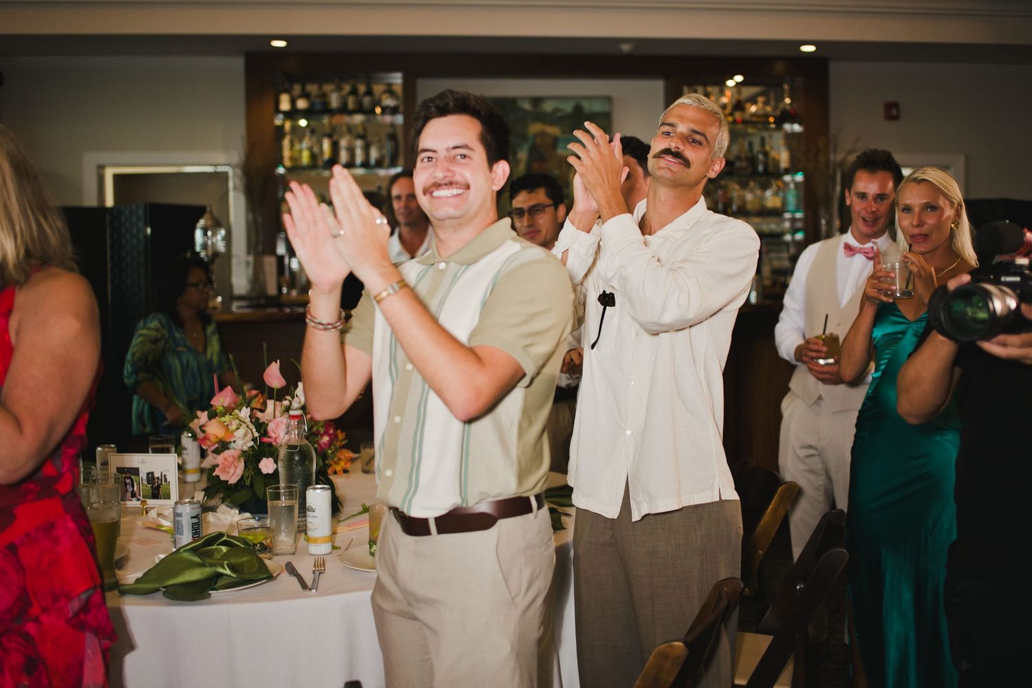 Guests clapping and smiling during a wedding reception at an elegant venue with a bar in the background.