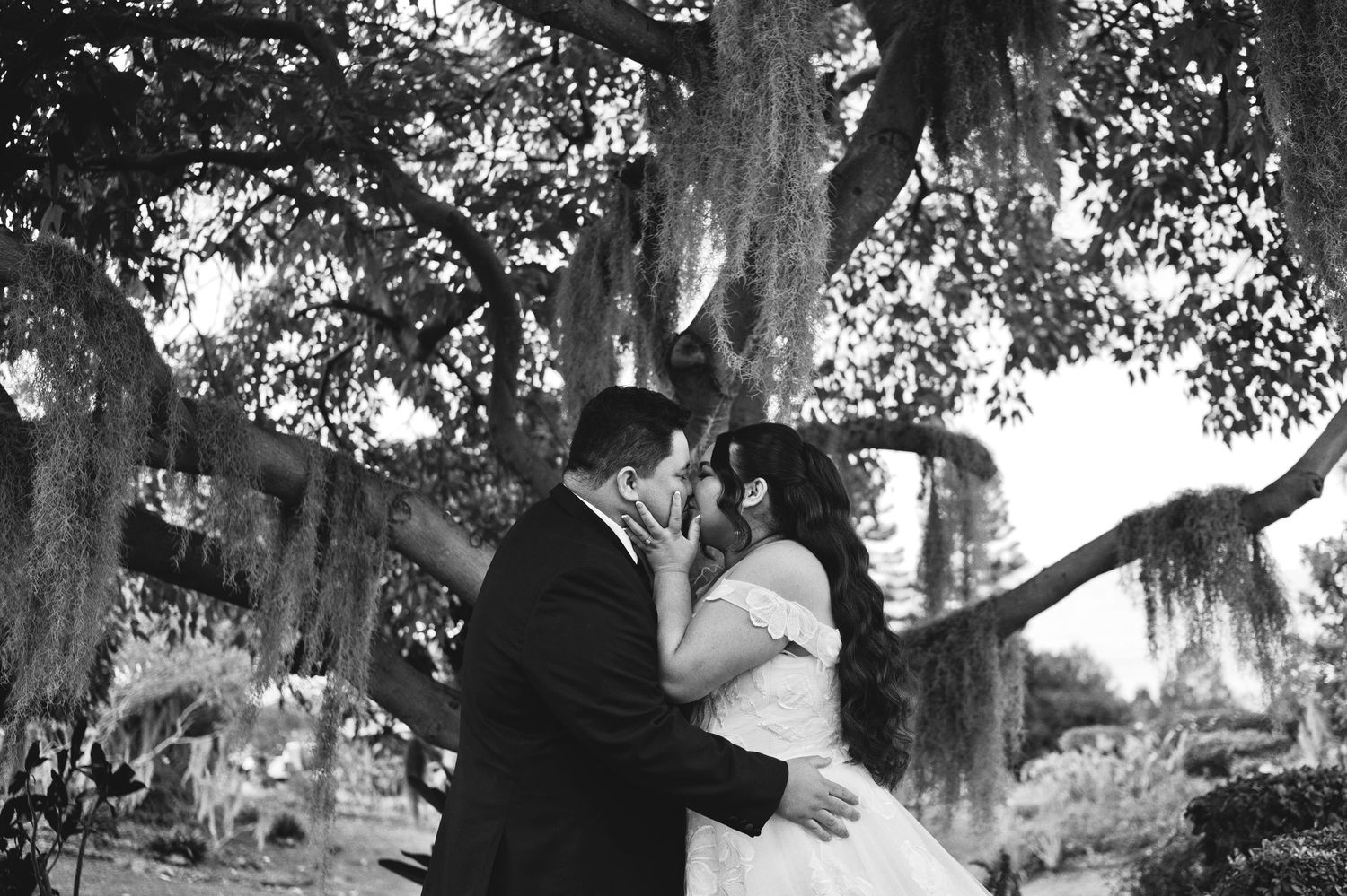 Black and white photo of a bride and groom sharing a romantic moment under a large moss-covered tree.