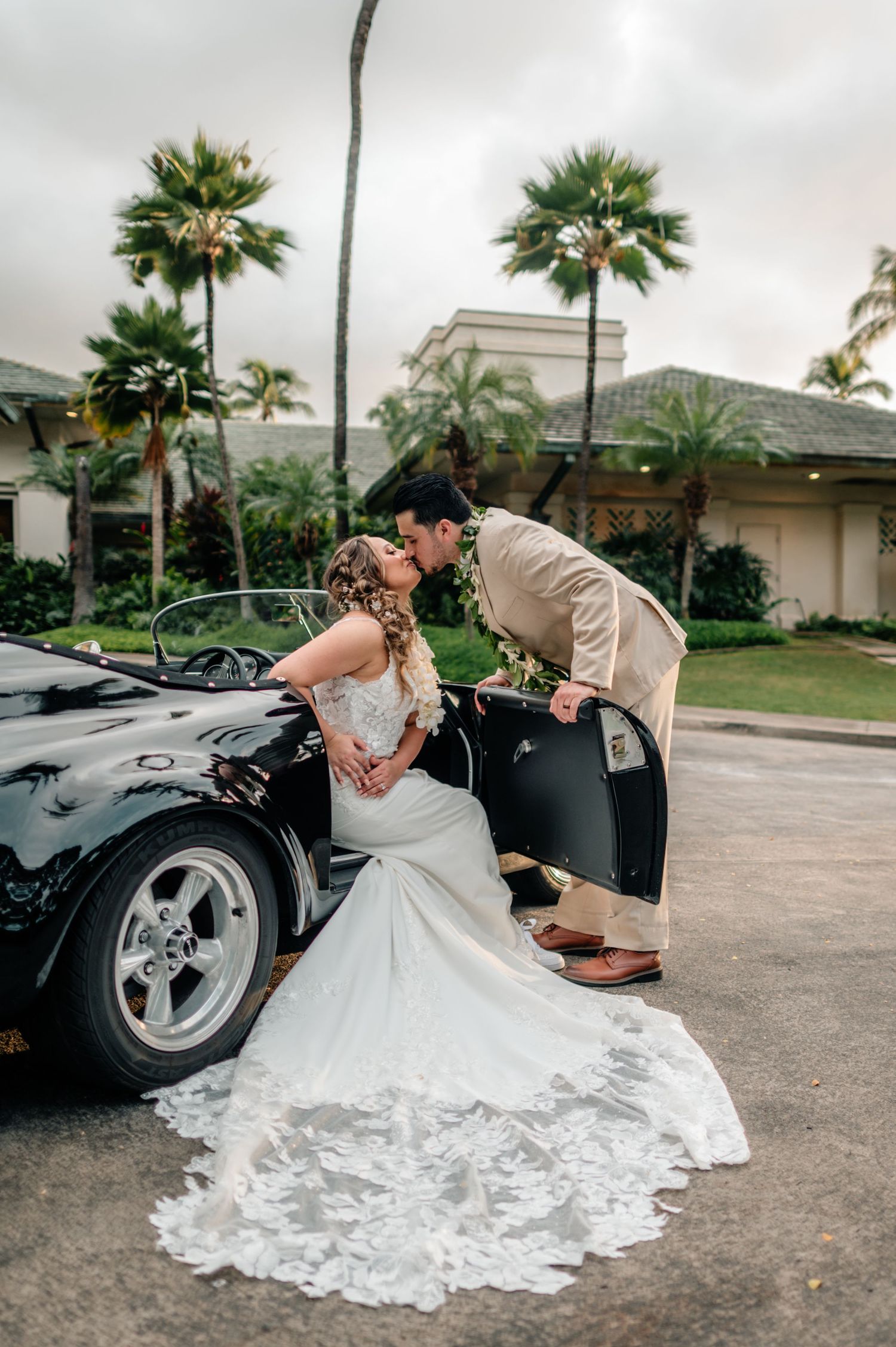 Bride and groom share a romantic kiss beside a classic black convertible sports car, with palm trees in the background.