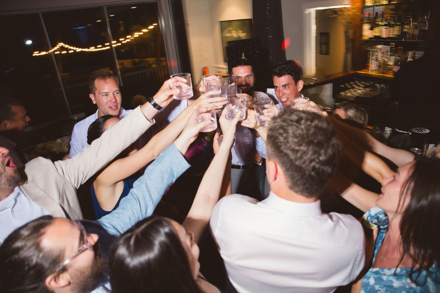 Wedding guests raise glasses in a joyful toast during a lively indoor wedding reception celebration.