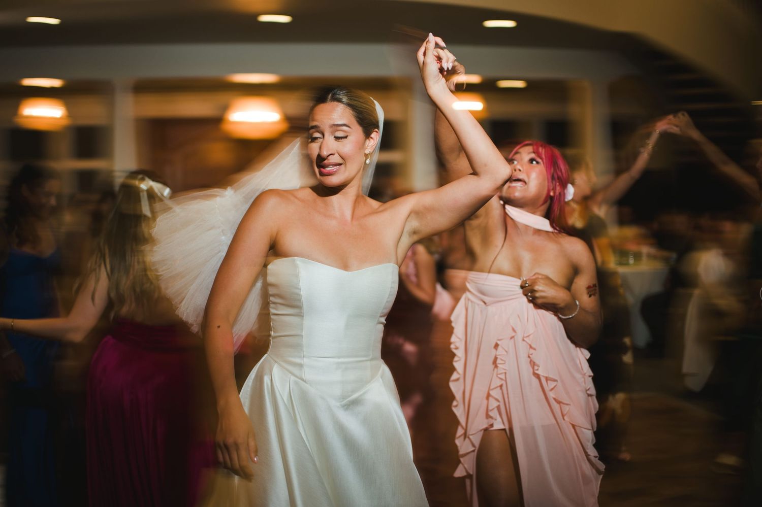 Bride in white strapless gown dancing joyfully with bridesmaids in pink dresses at a wedding reception.