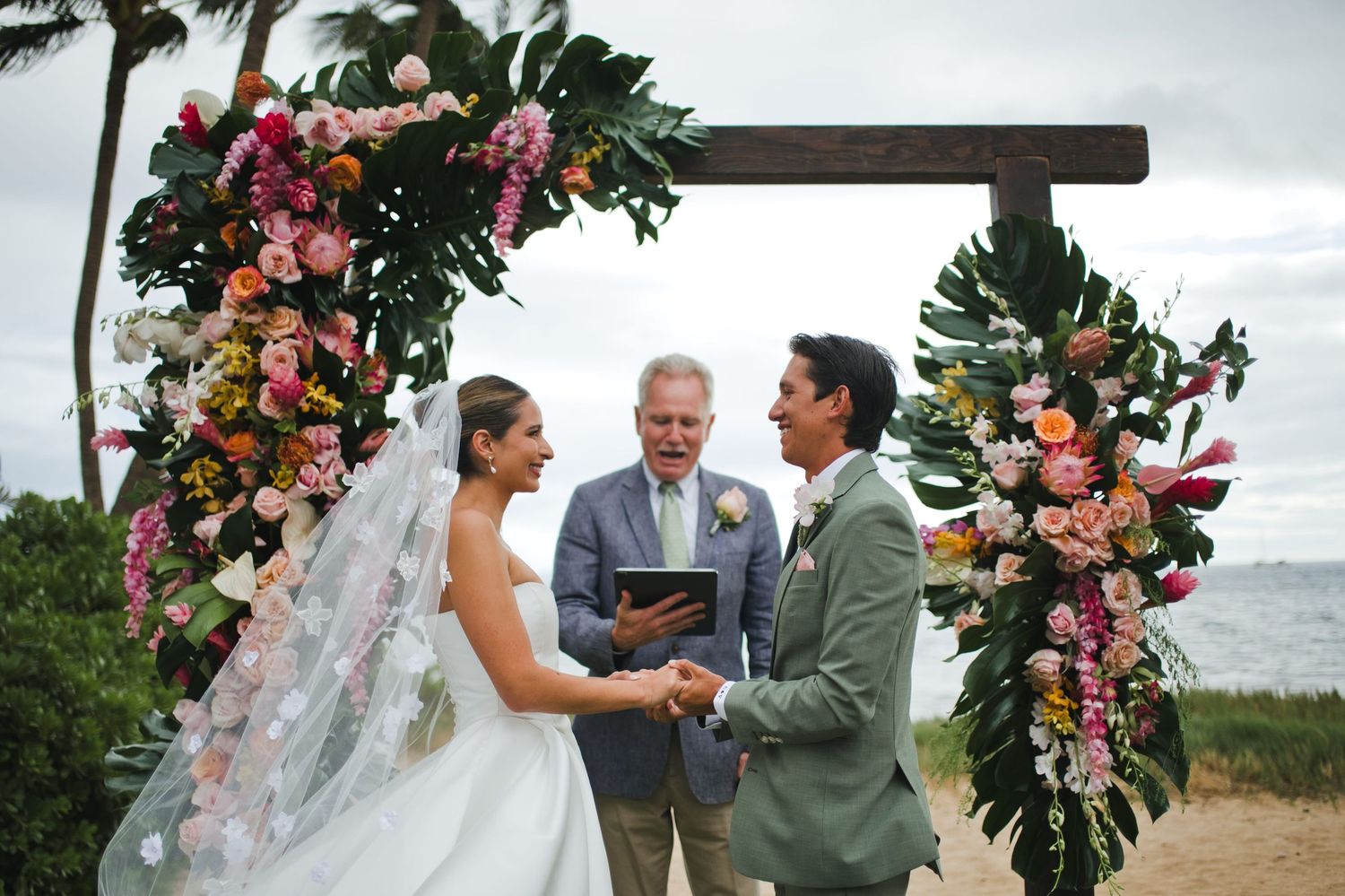 Bride and groom exchange vows under a floral arch with tropical flowers at an outdoor beach wedding ceremony.