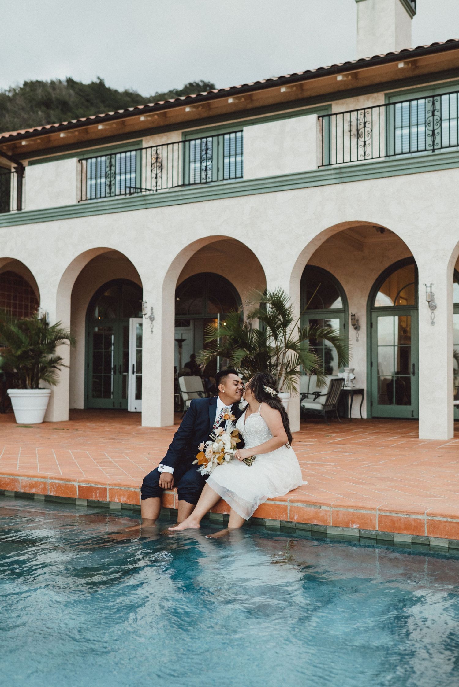 A couple in formal attire playfully falls into a swimming pool at an elegant Spanish-style venue.