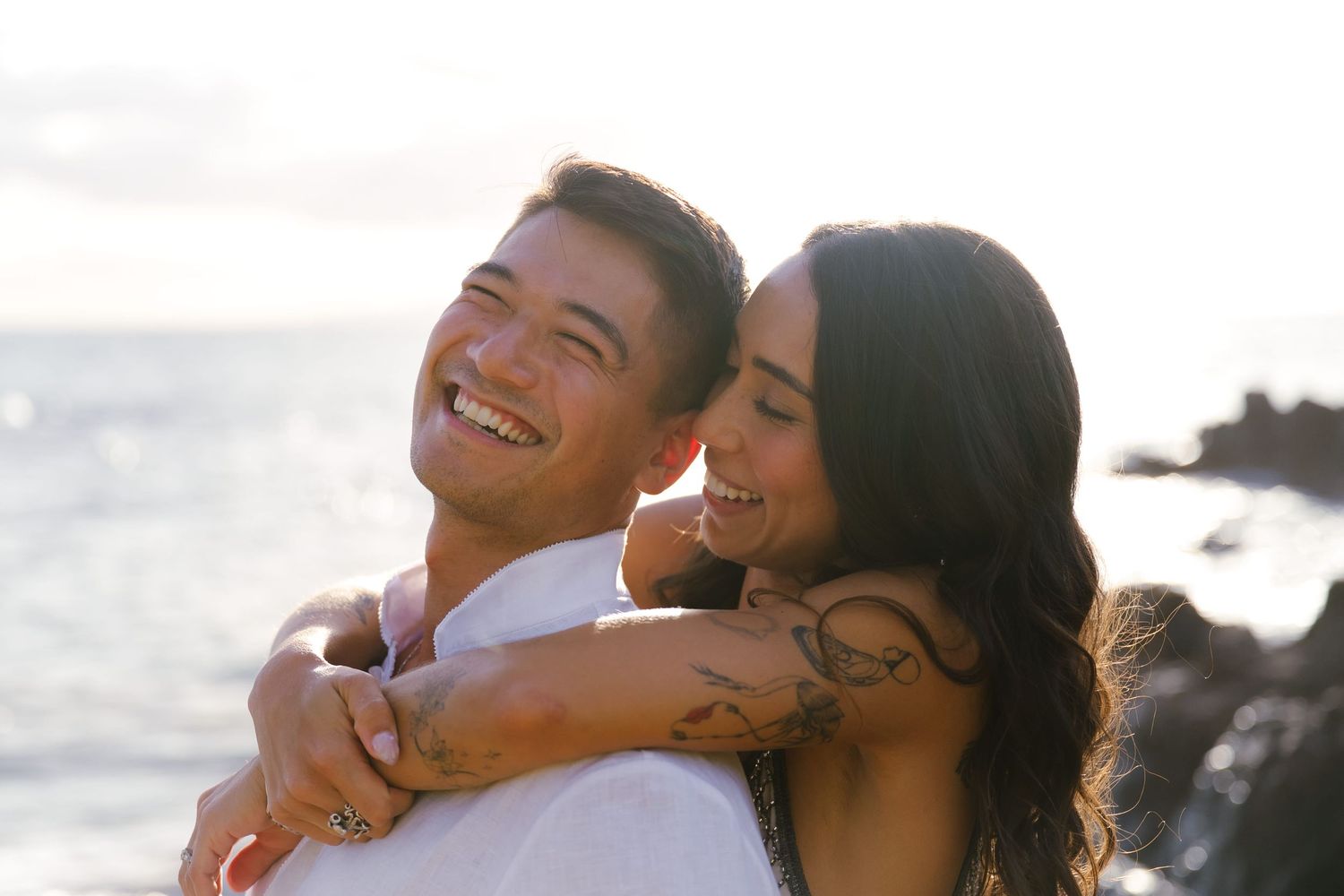 Couple embracing and smiling at sunset by the ocean with tattoo visible on arm.
