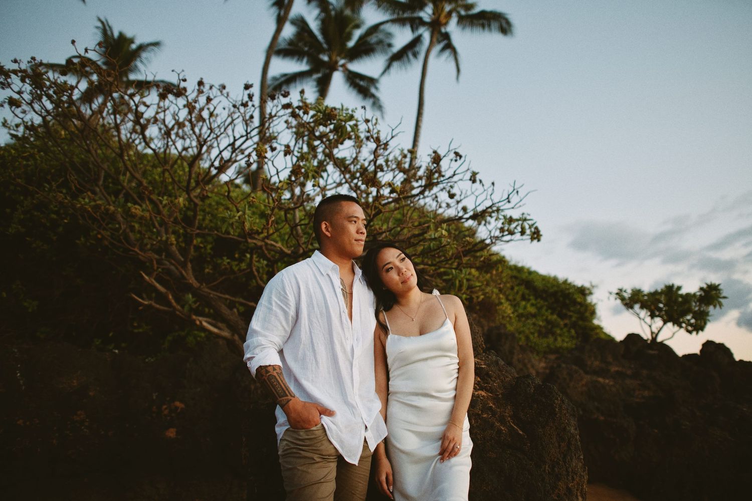 Couple leaning together at dusk near tropical palms and lava rocks in Maui Hawaii.
