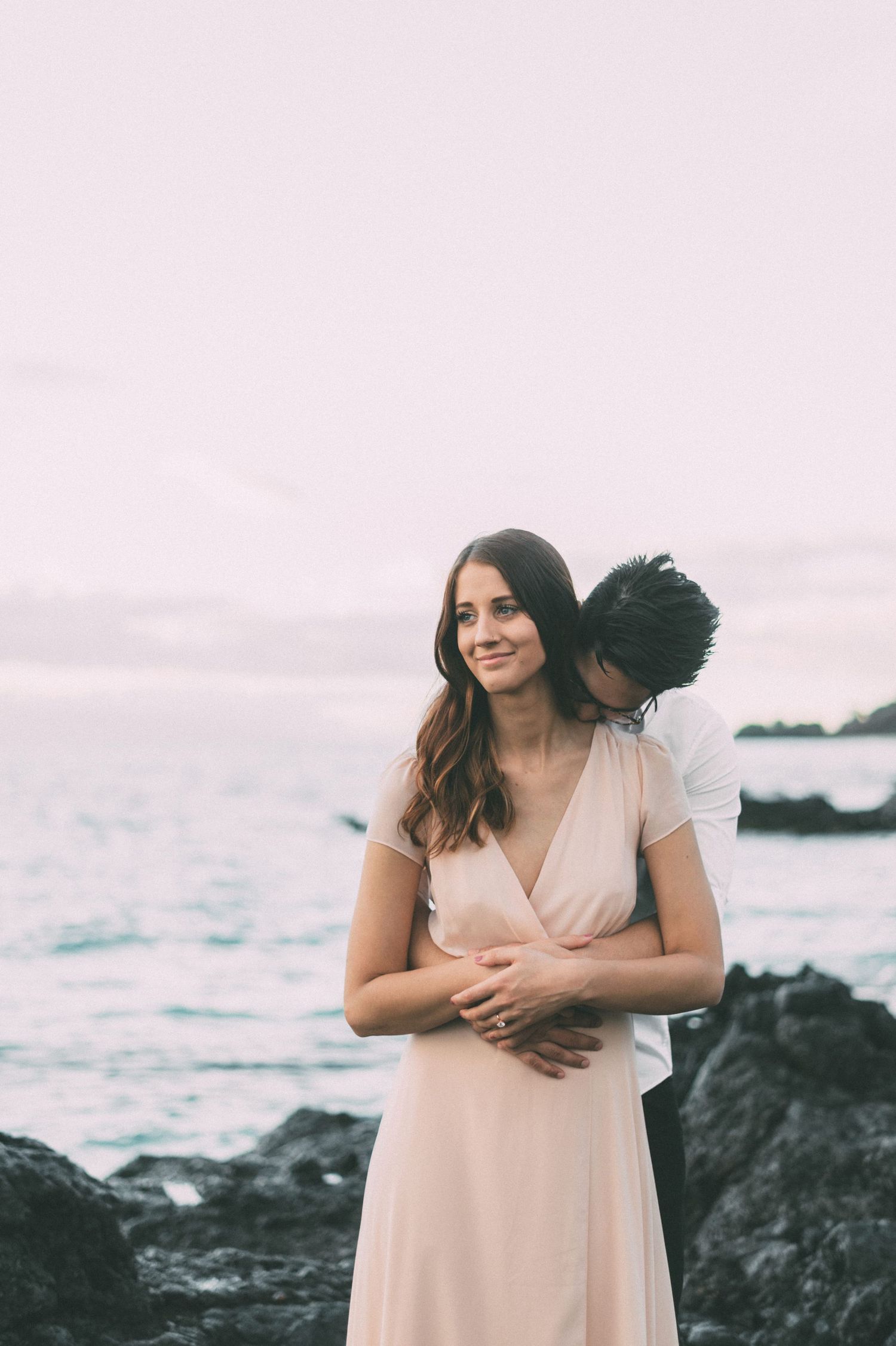 Woman in blush pink dress smiling as partner embraces her from behind near ocean rocks.