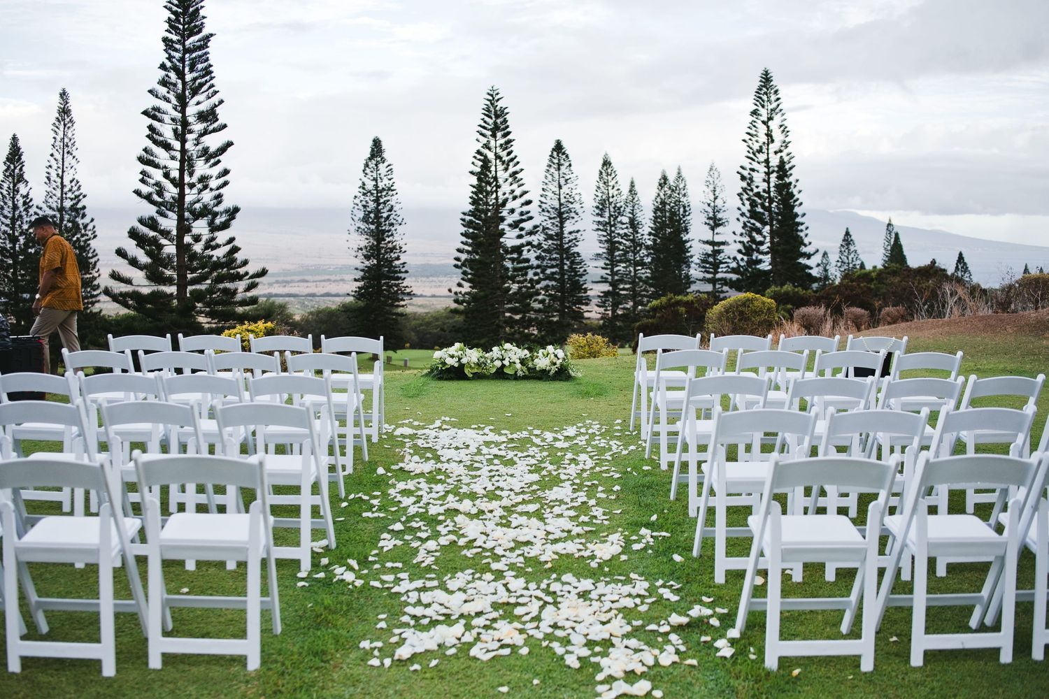 Outdoor wedding ceremony setup with white chairs, flower petal aisle, and tall pine trees on a lush green lawn.