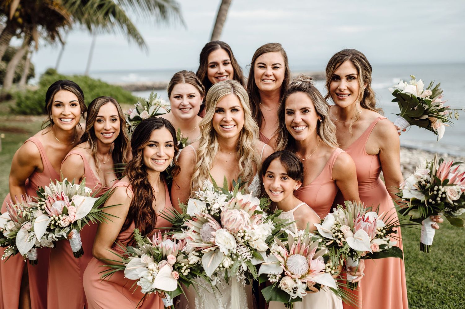 Bride and bridesmaids in coral dresses holding tropical bouquets with king protea flowers at an oceanside wedding.