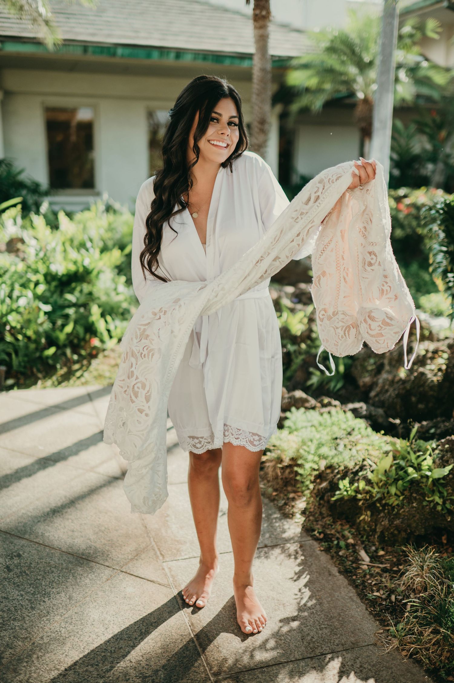 Smiling bride in white robe holding lace overlay outdoors in tropical garden setting.