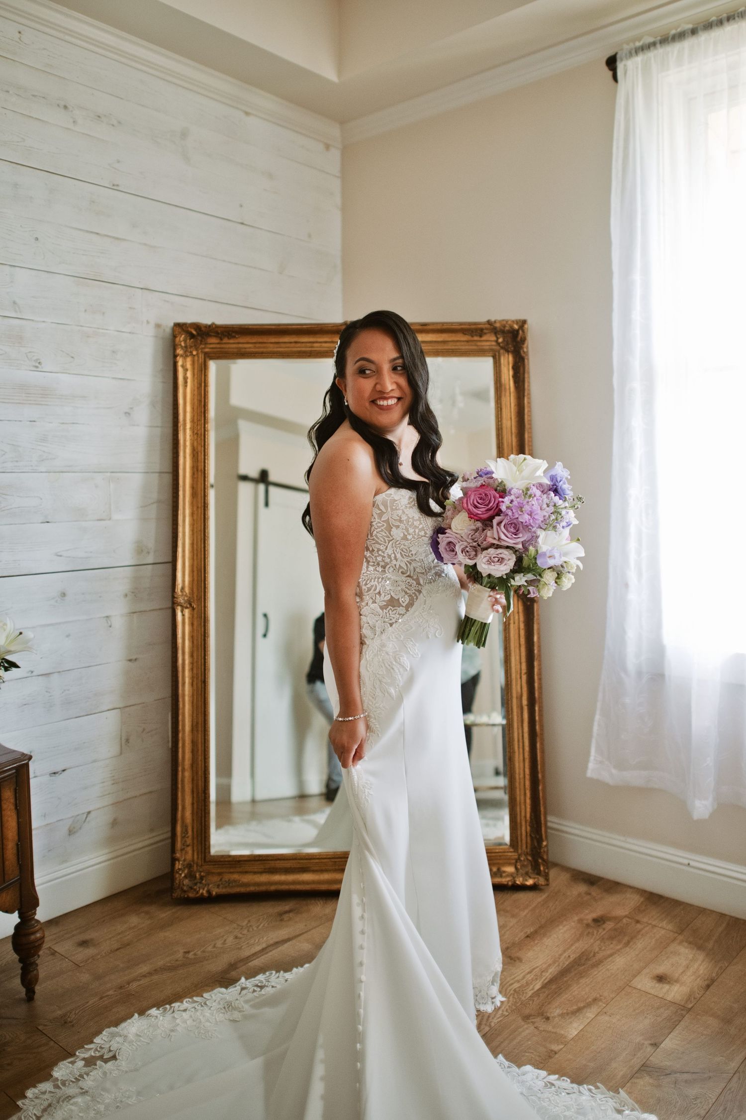 Bride in white lace gown holding purple bouquet stands before gold mirror in bright room.