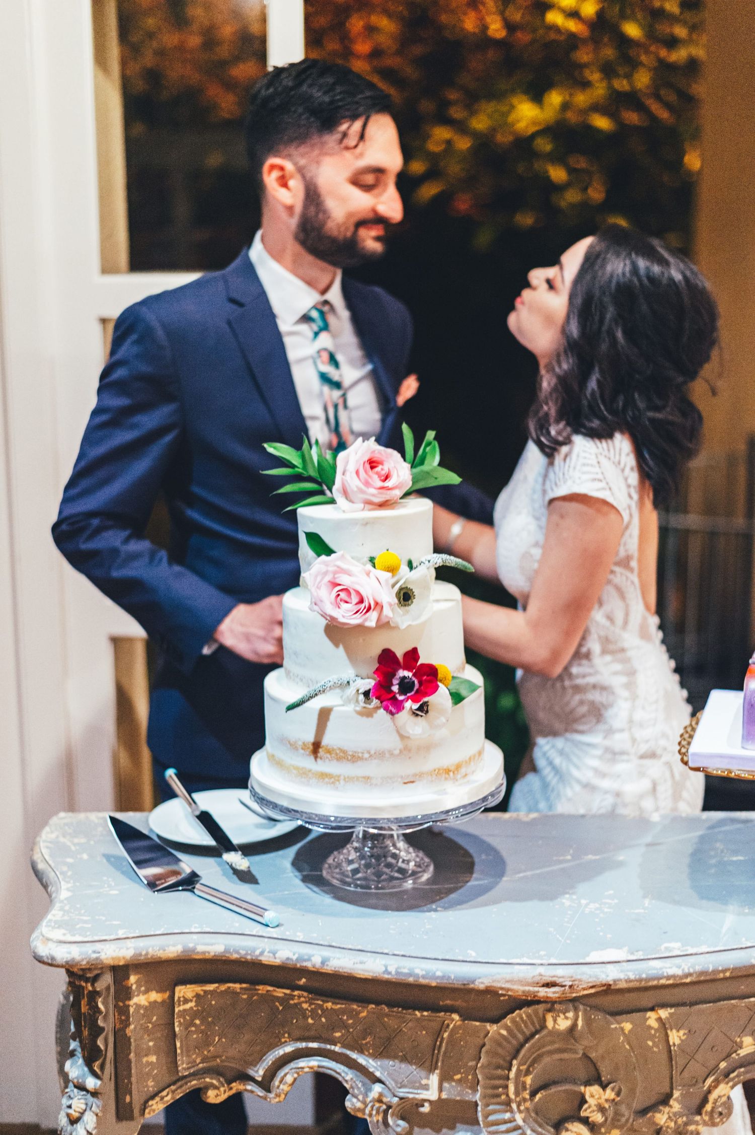 Couple cutting a floral three-tier white wedding cake decorated with pink roses and anemones on ornate table.
