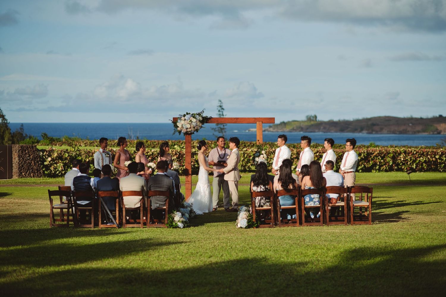 Outdoor wedding ceremony on lush green lawn with ocean backdrop, wooden arch, and guests seated in wooden chairs.