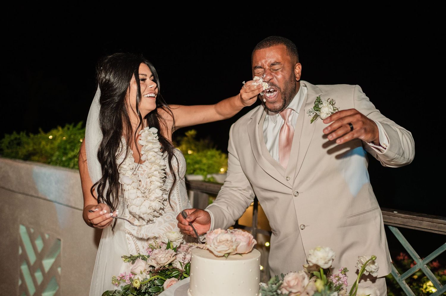 Bride feeds wedding cake to groom during outdoor nighttime reception, both dressed in elegant wedding attire.
