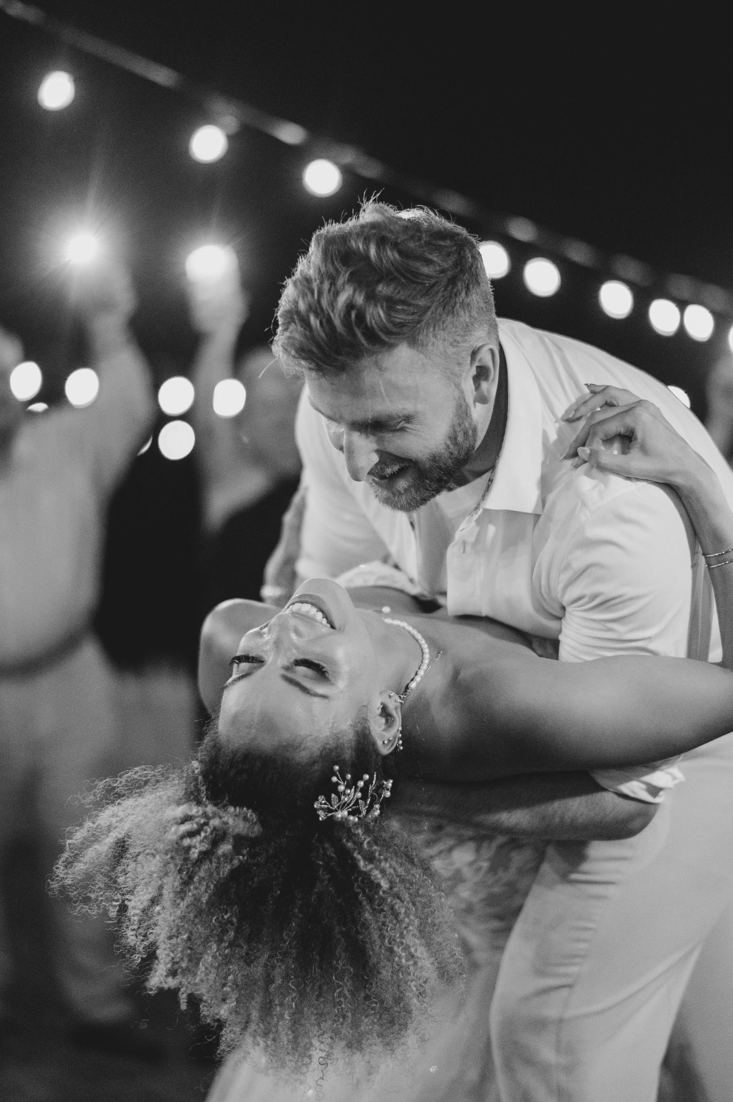 Black and white photo of a couple sharing a romantic dip dance under string lights at a wedding reception.