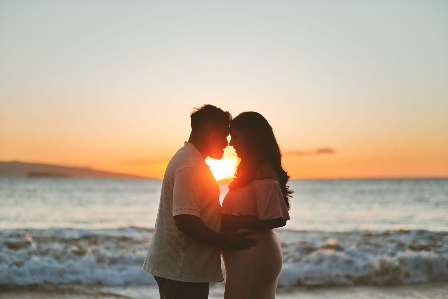 Expecting parents sharing a tender moment on a beach at sunset with golden light glowing between them.