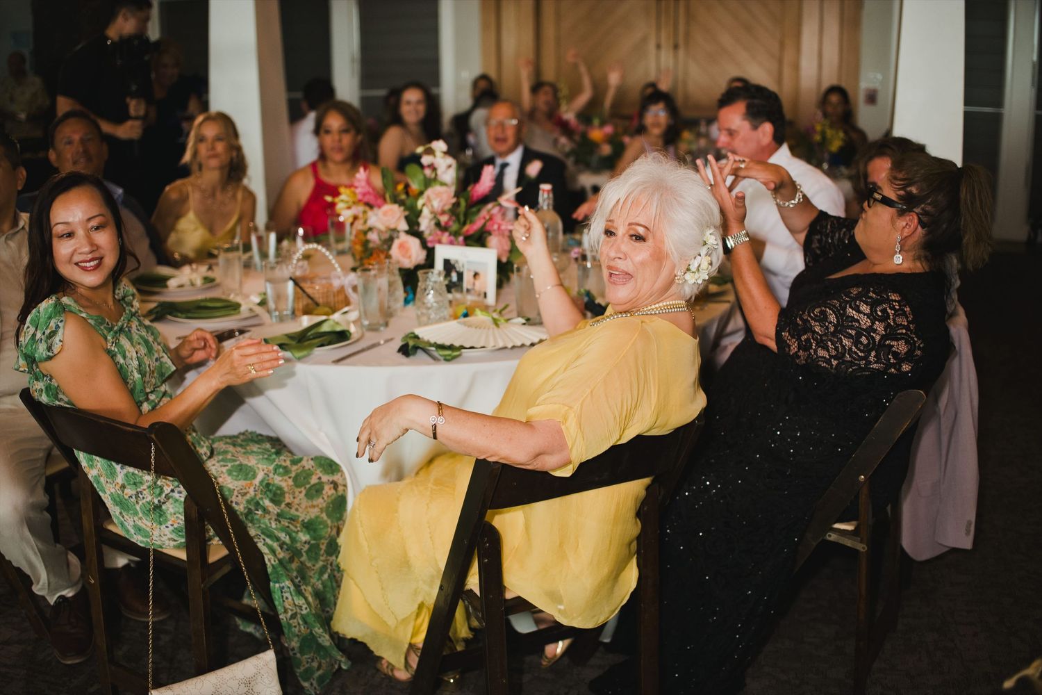 Guests laughing and celebrating at a wedding reception dinner, wearing elegant attire at decorated tables.