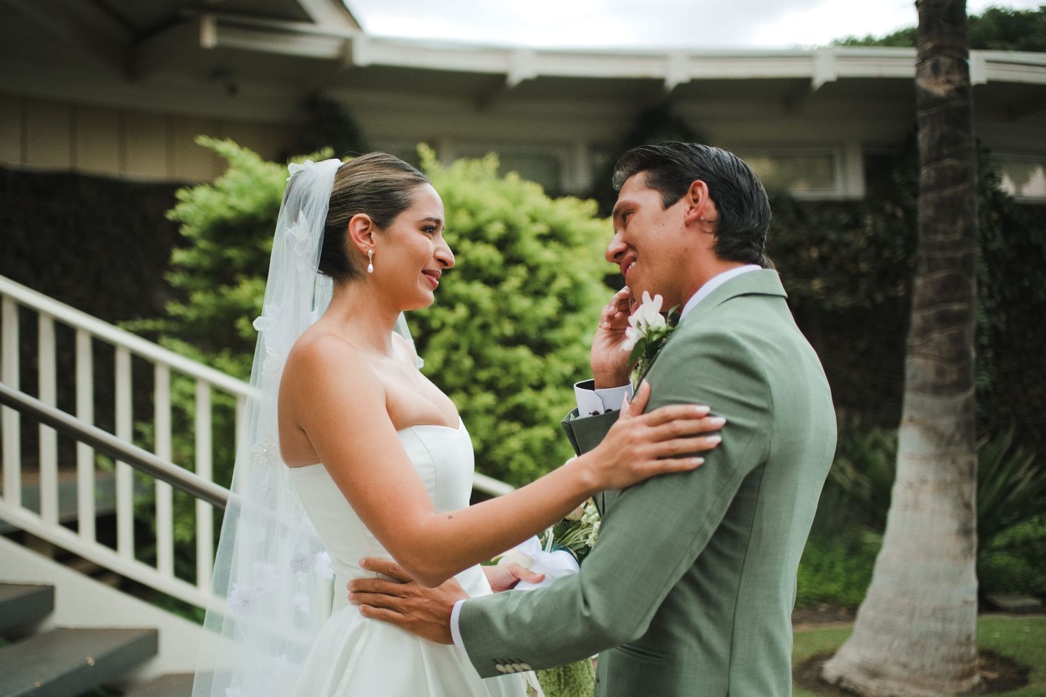 Bride and groom share an emotional first look moment outdoors, with the bride in a white gown and veil and groom in a sage suit.