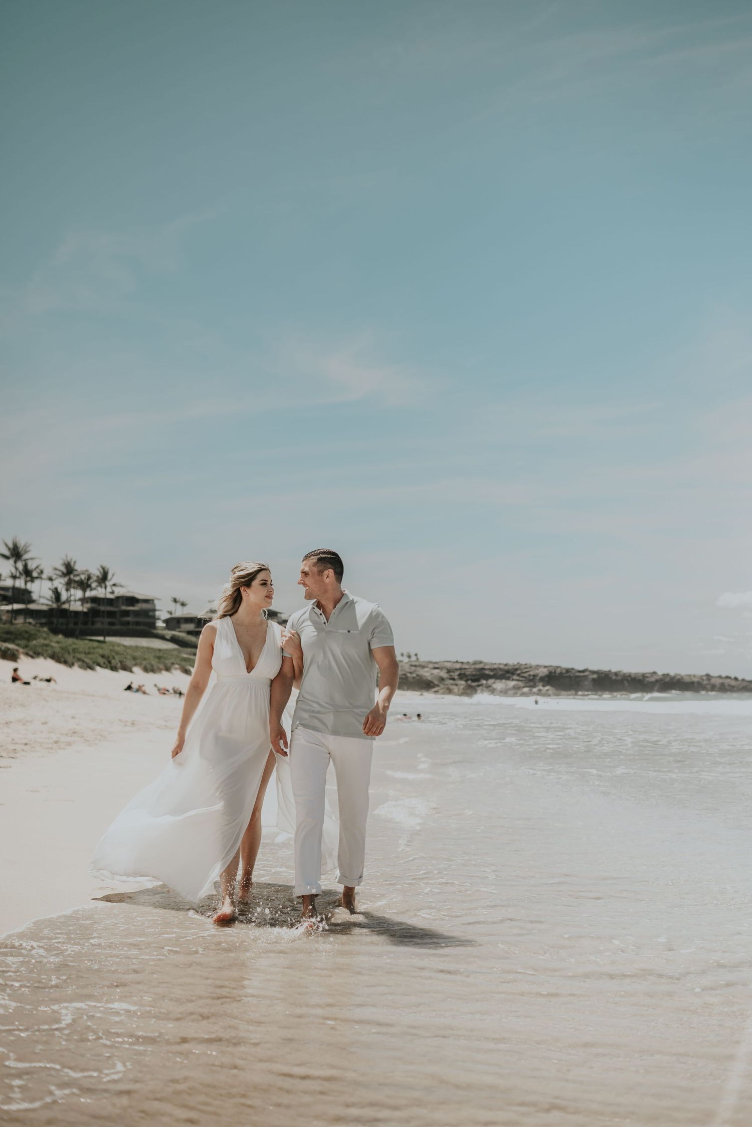 Couple in white outfits walking along a sunny beach shoreline smiling at each other.