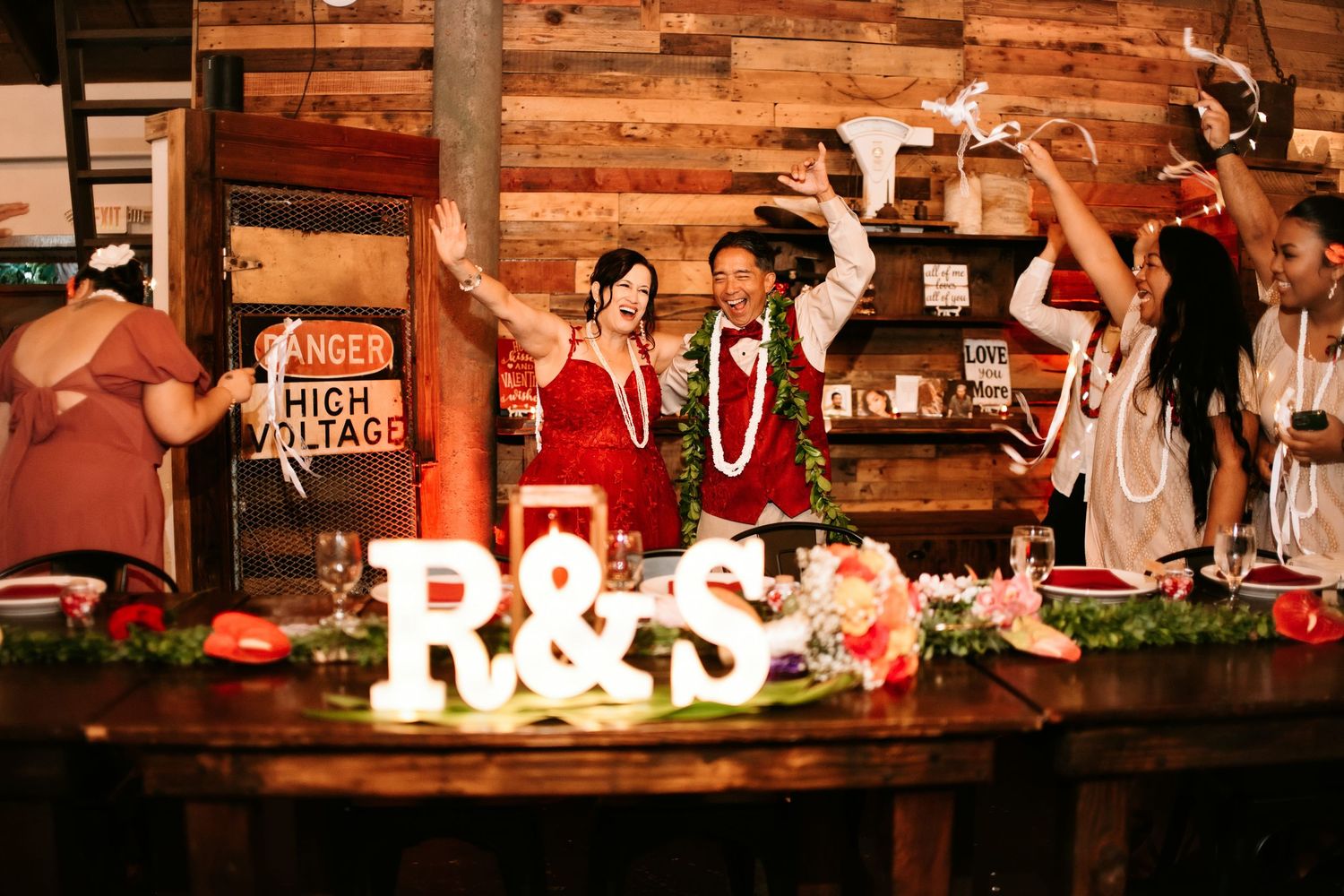 Couple celebrates at wedding sweetheart table with R&S marquee letters, surrounded by guests in a rustic barn venue.