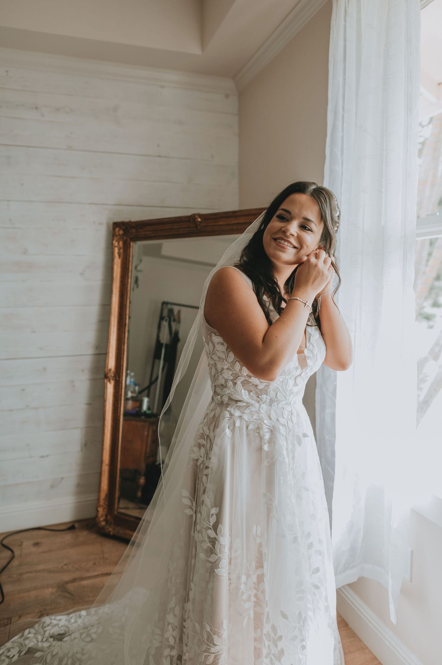 Smiling bride in lace wedding gown and veil adjusting earring by window with ornate gold mirror.