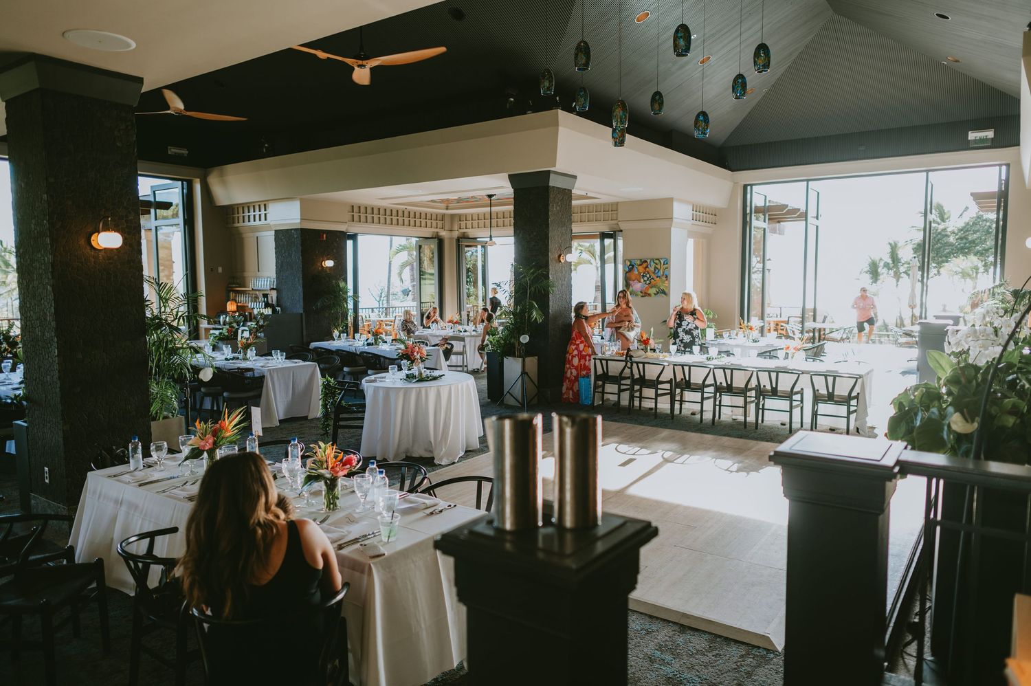 Elegant wedding reception venue with white-draped tables, tropical floral arrangements, and floor-to-ceiling windows overlooking a sunny out