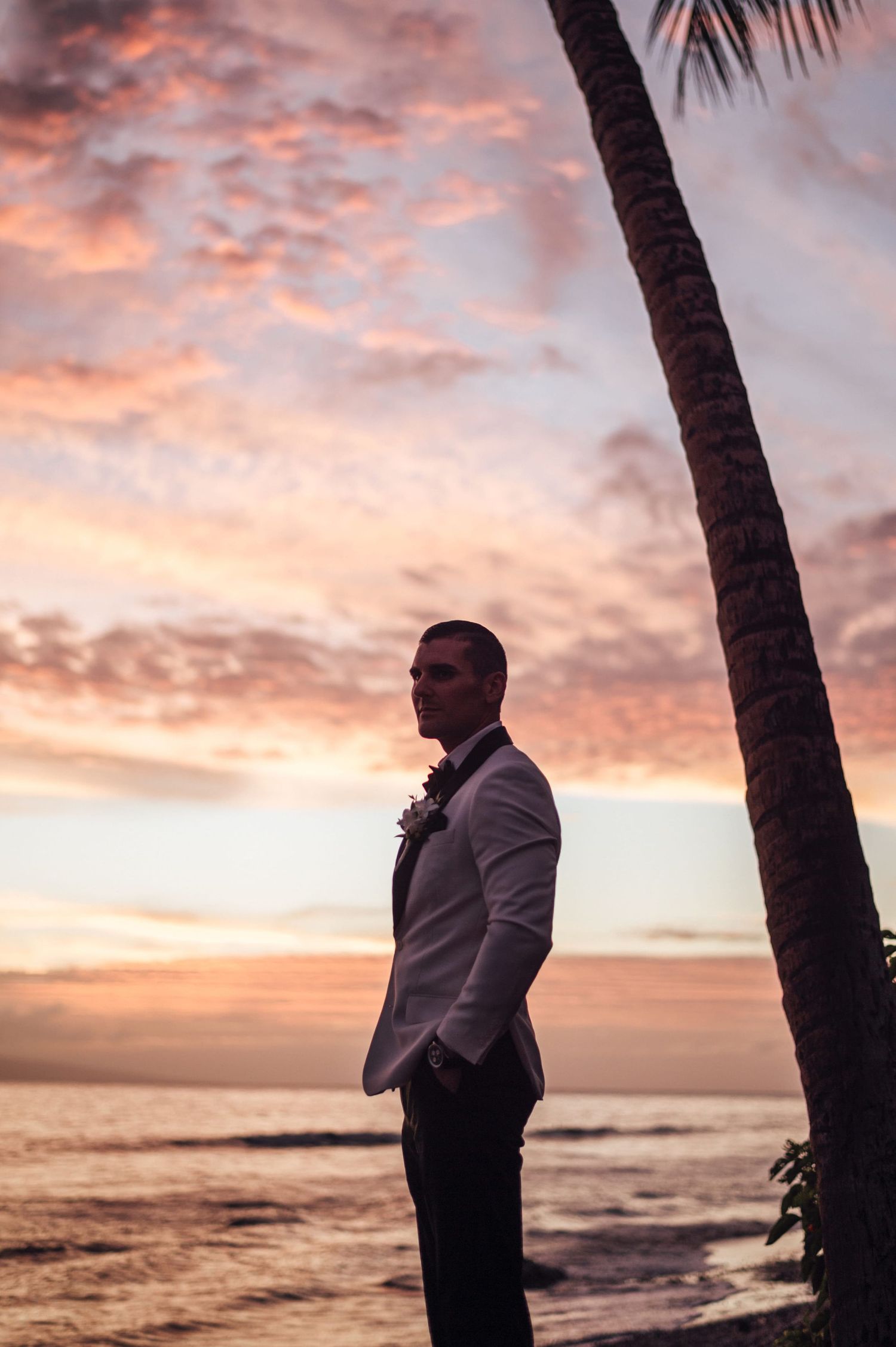 Groom in white blazer stands by palm tree at tropical beach sunset during wedding celebration.