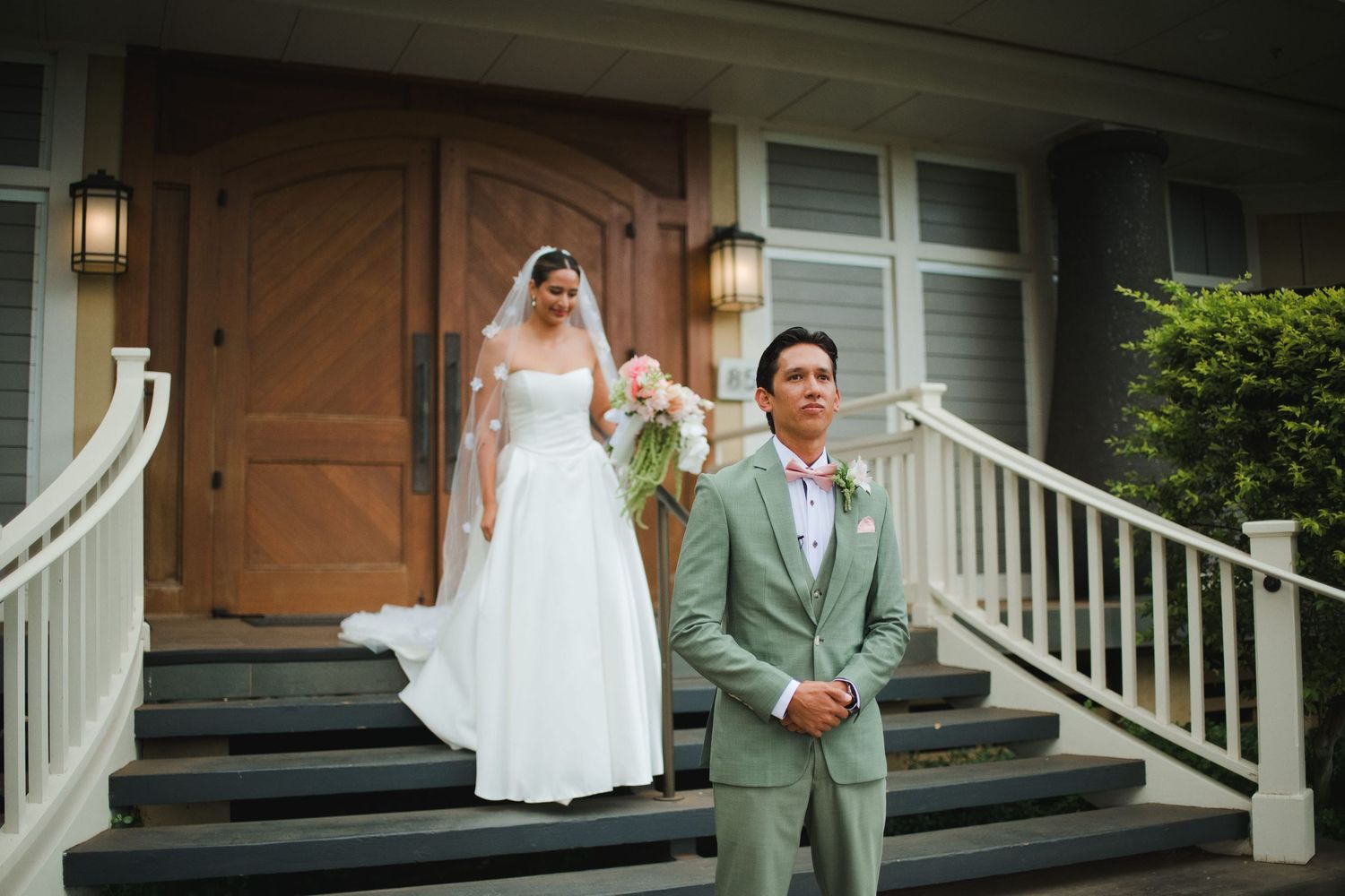 Bride in white gown stands on steps behind groom in sage suit during romantic outdoor first look wedding moment.