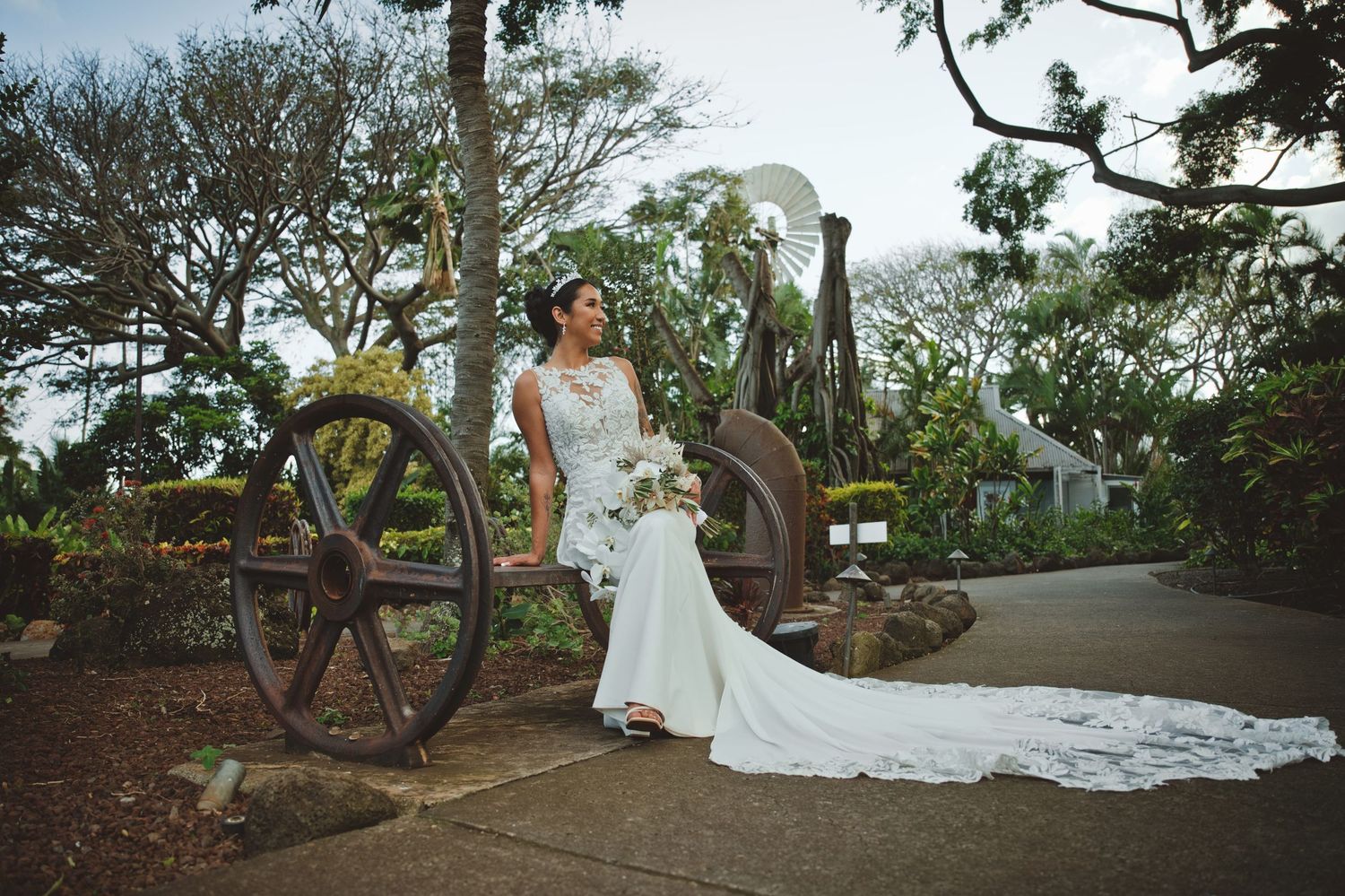 Bride in white lace gown with long train poses beside rustic antique wagon wheel in lush garden setting.