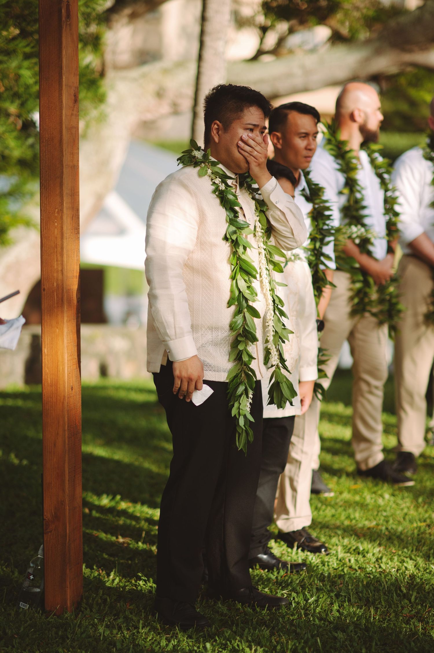 Emotional groom wearing a green leaf lei wipes tears at an outdoor Hawaiian wedding ceremony with groomsmen.