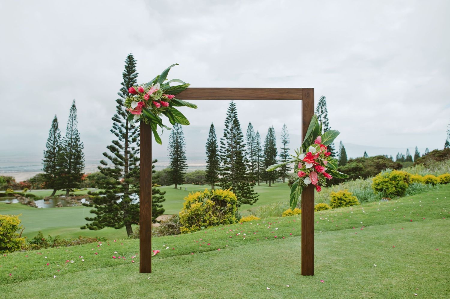 Wooden wedding arch adorned with tropical pink floral arrangements on a lush green lawn with pine trees backdrop.