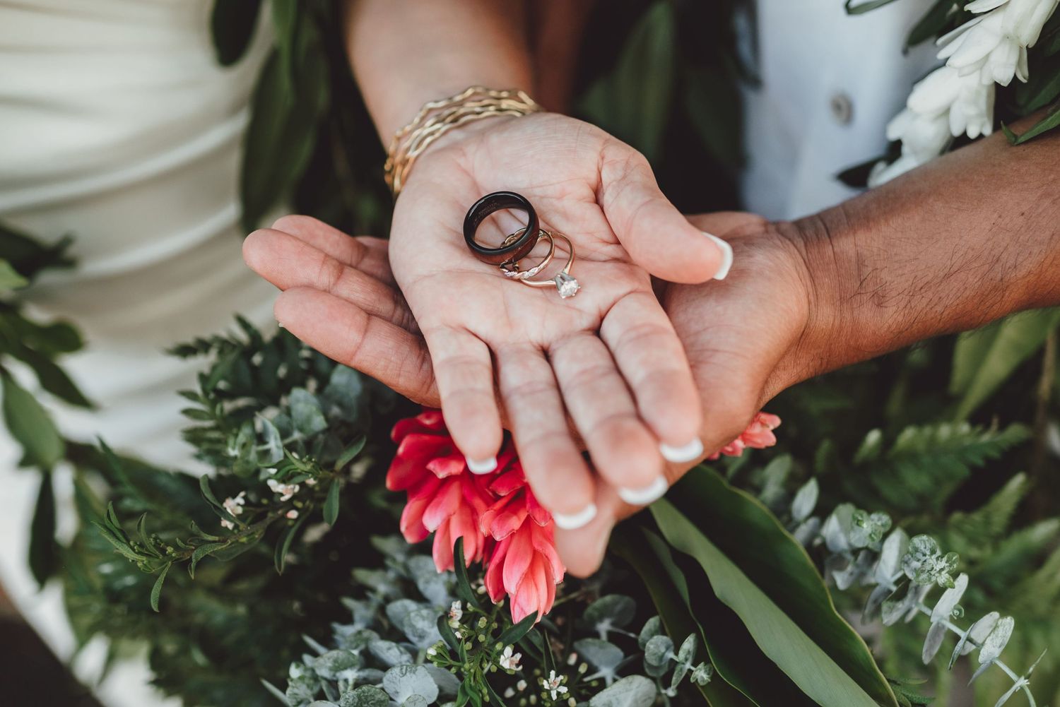 Two hands holding wedding rings over a floral bouquet with pink flowers and greenery.