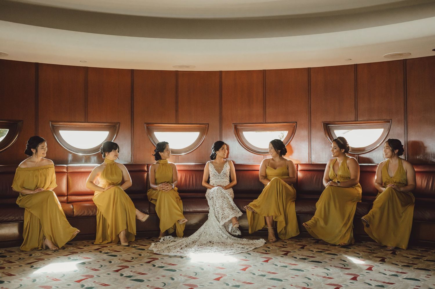 Bride in white gown sits with six bridesmaids in yellow dresses on curved bench inside elegant venue.