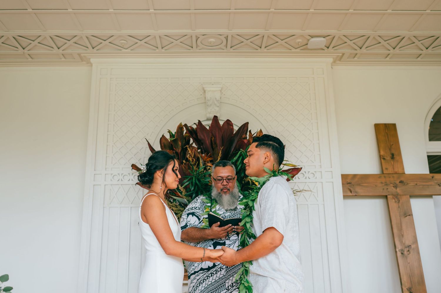 Couple exchanges vows before officiant at tropical floral altar in white chapel with wooden cross.