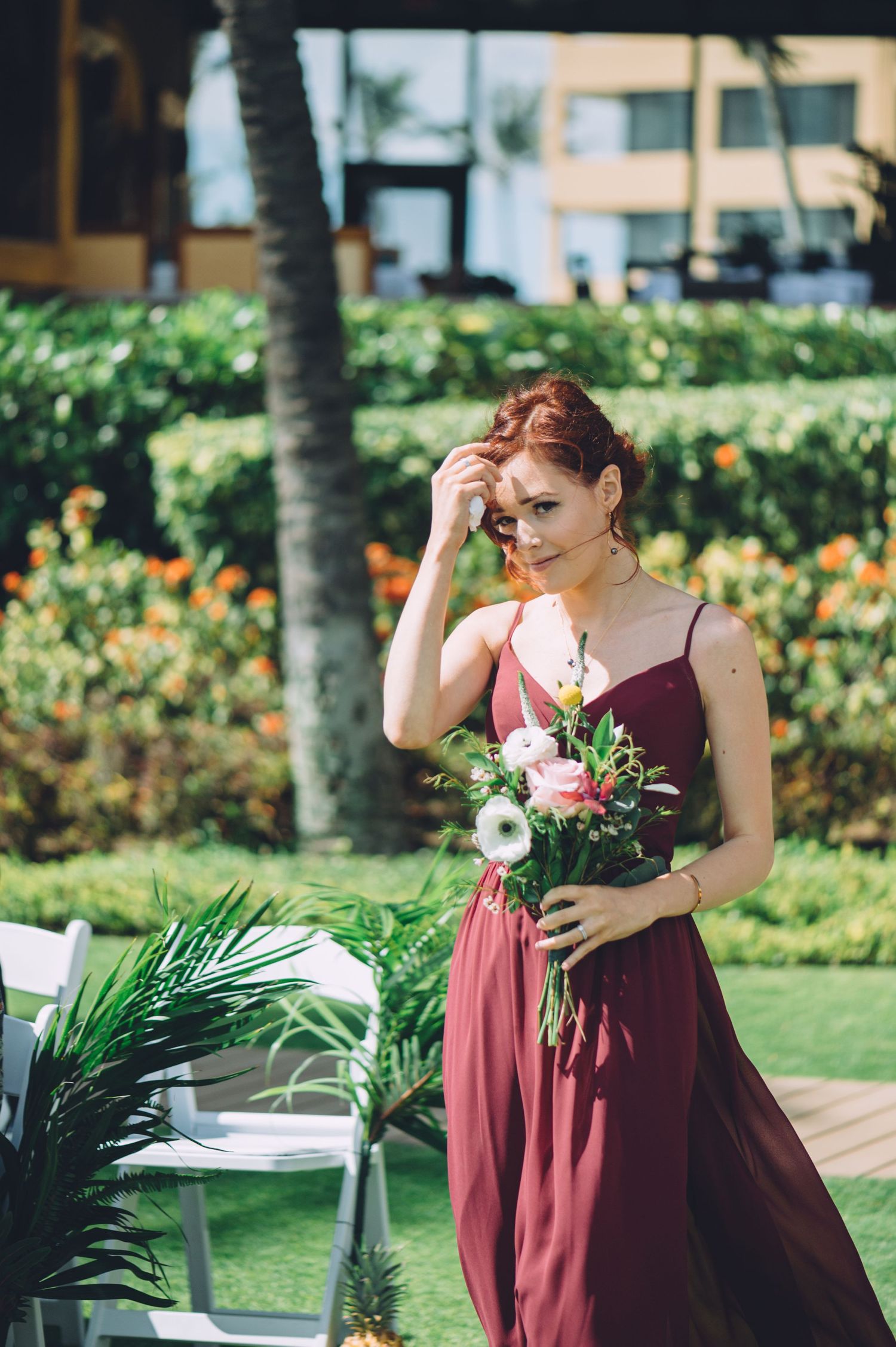 Bridesmaids in burgundy dresses hold bouquets during an outdoor garden ceremony.