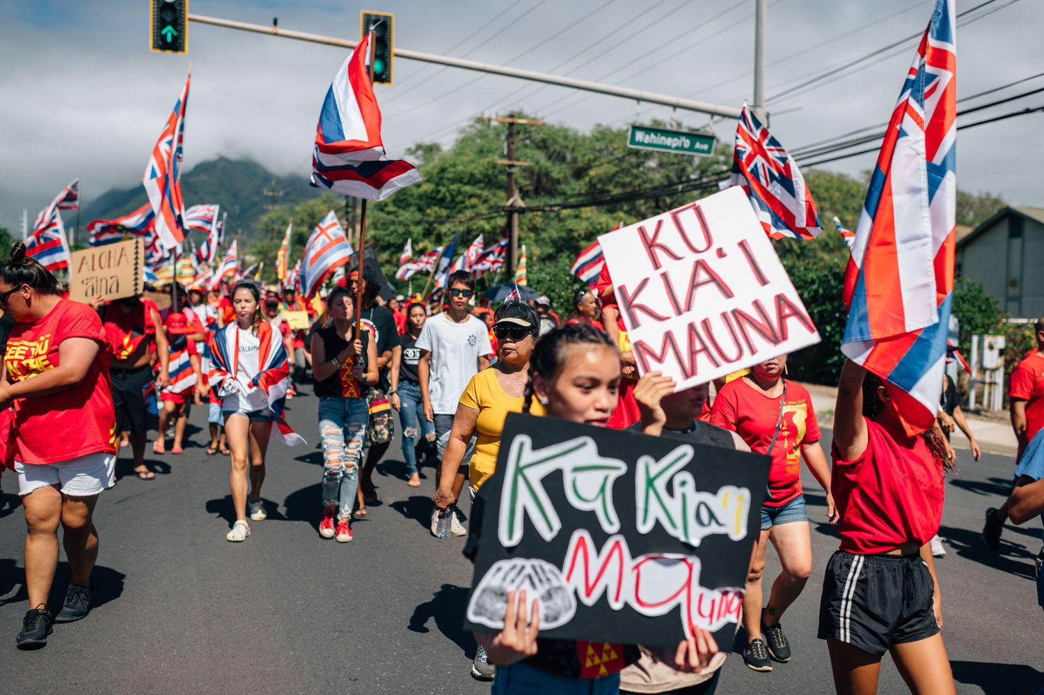 Crowd marches with protest signs and Hawaiian flags through street intersection.