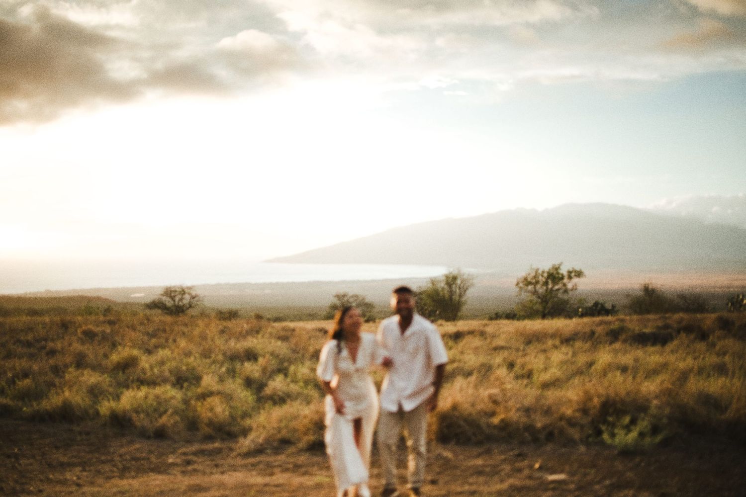 Couple walking hand in hand through golden Hawaiian fields at sunset with ocean and mountains in background.