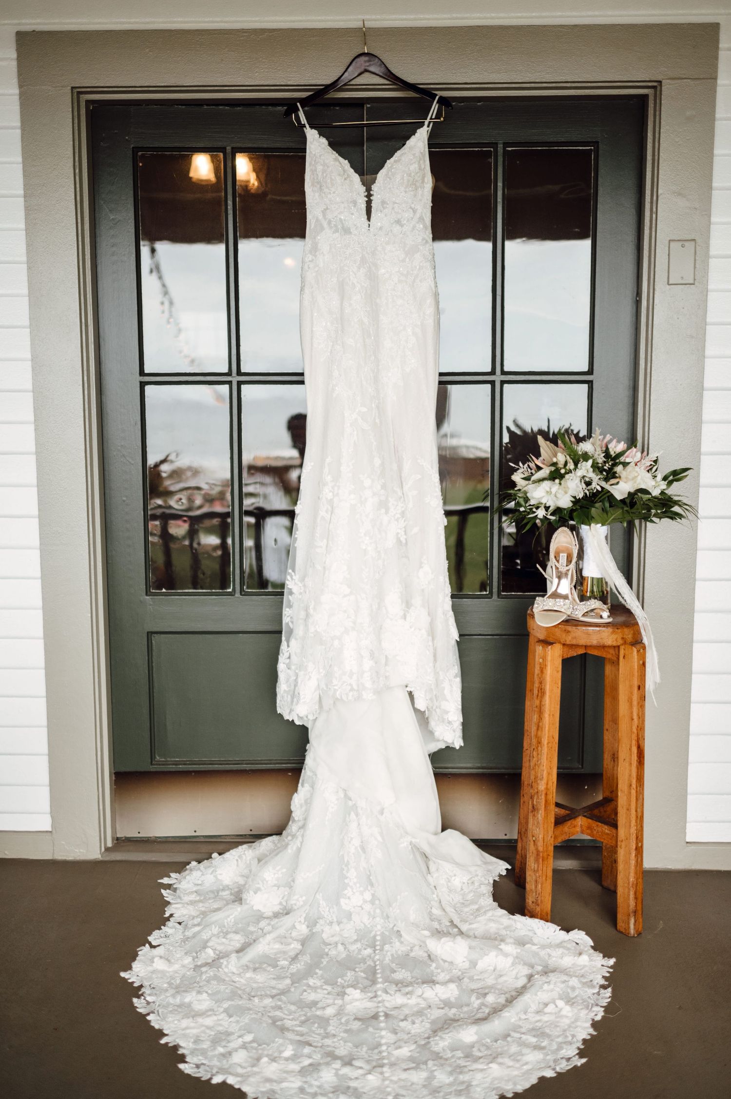 White lace wedding dress with train hanging on a green door beside a wooden stool with bridal bouquet and heels.