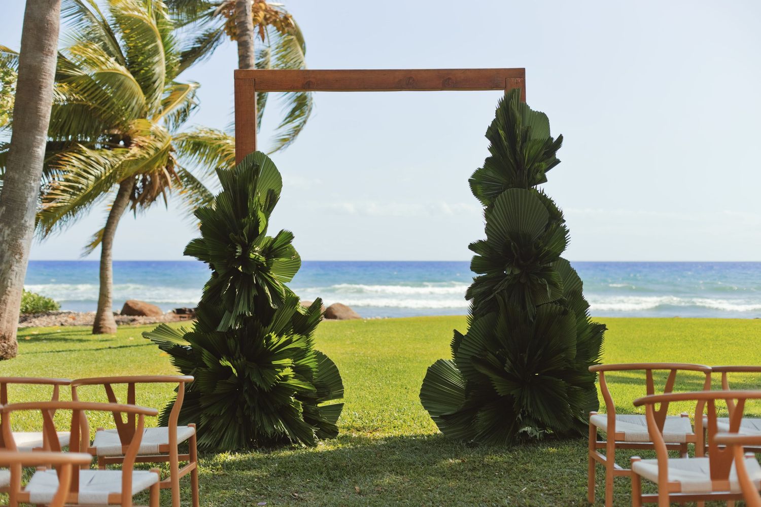Tropical beach wedding arch adorned with lush green leaves, set on oceanfront lawn with palm trees and turquoise sea.