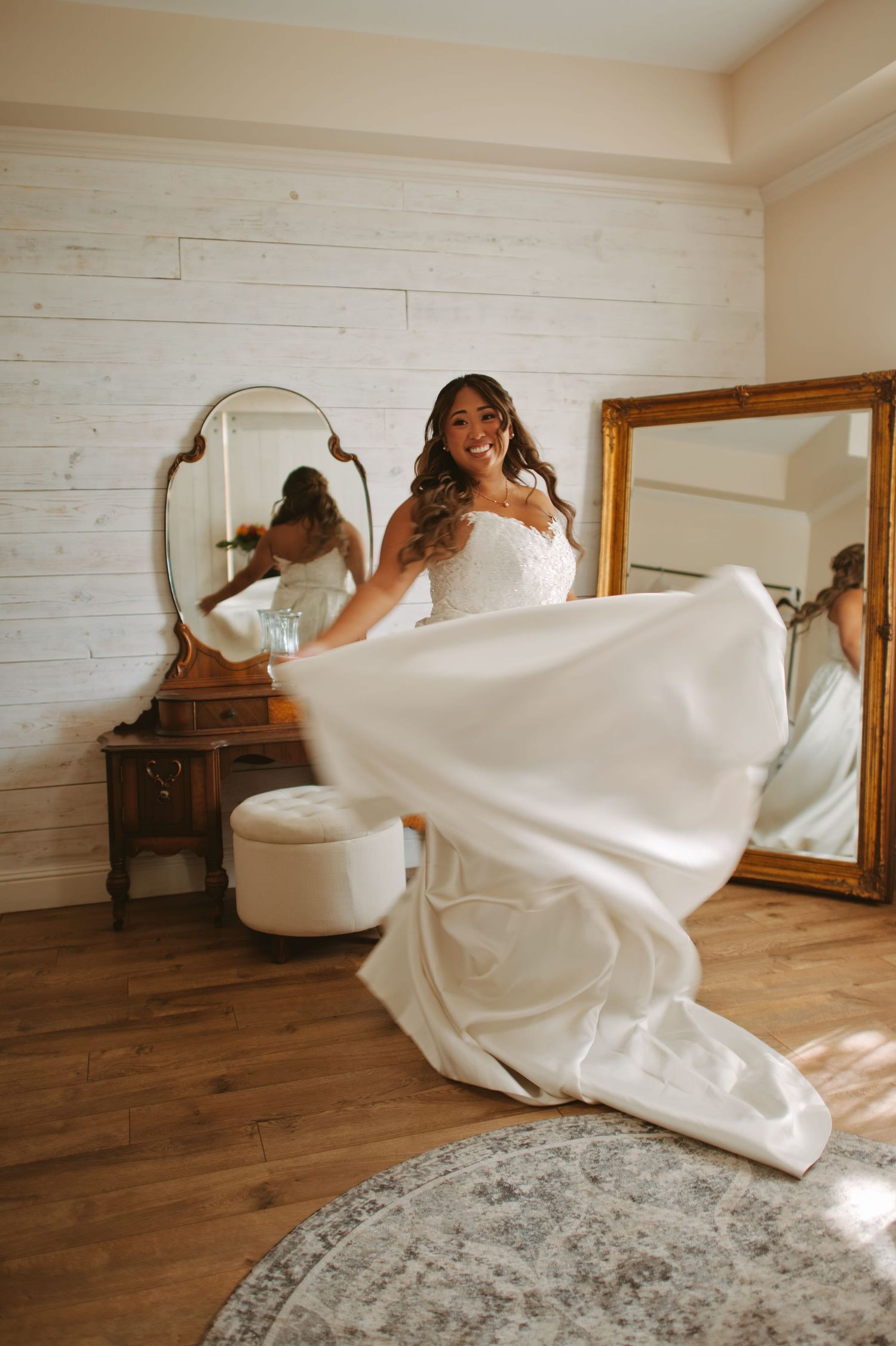 Smiling bride in white wedding gown twirling in a room with vintage mirrors and hardwood floors.