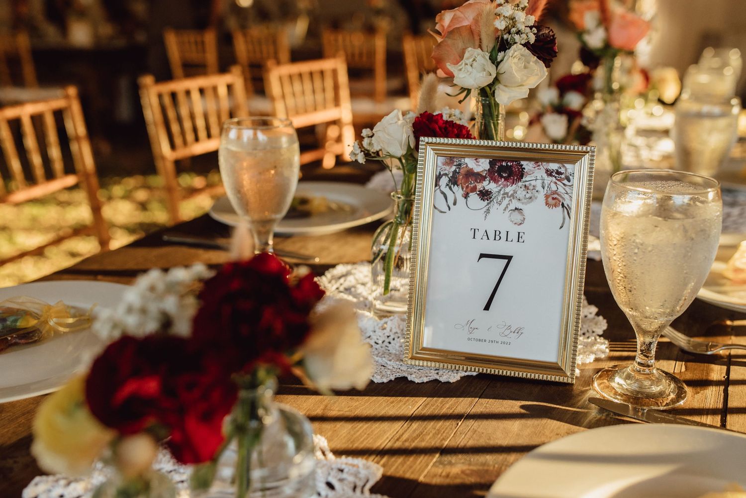Elegant wedding reception table setting with Table 7 sign, red and white floral centerpieces, and gold chiavari chairs.