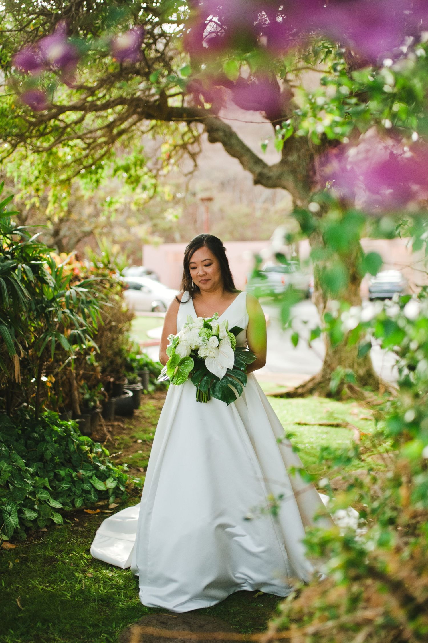 Bride in white ball gown holding white bouquet stands in lush garden path framed by purple flowering trees.