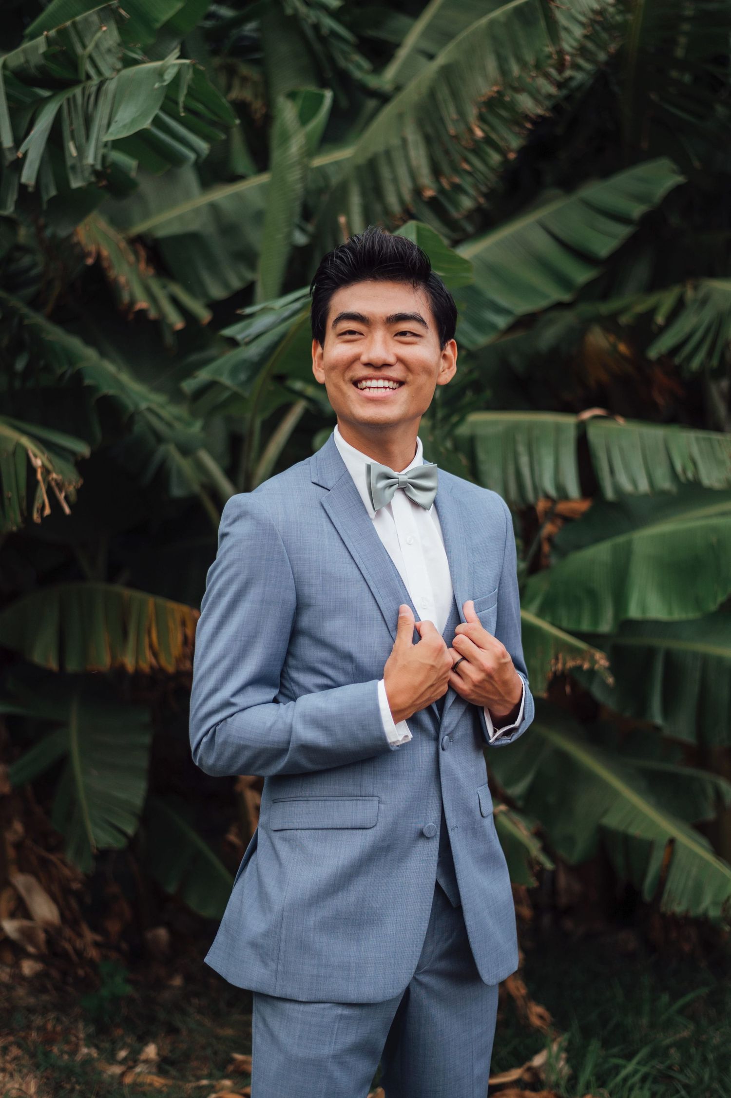 Smiling groom in light blue suit and sage bow tie posing outdoors in front of tropical banana leaves.
