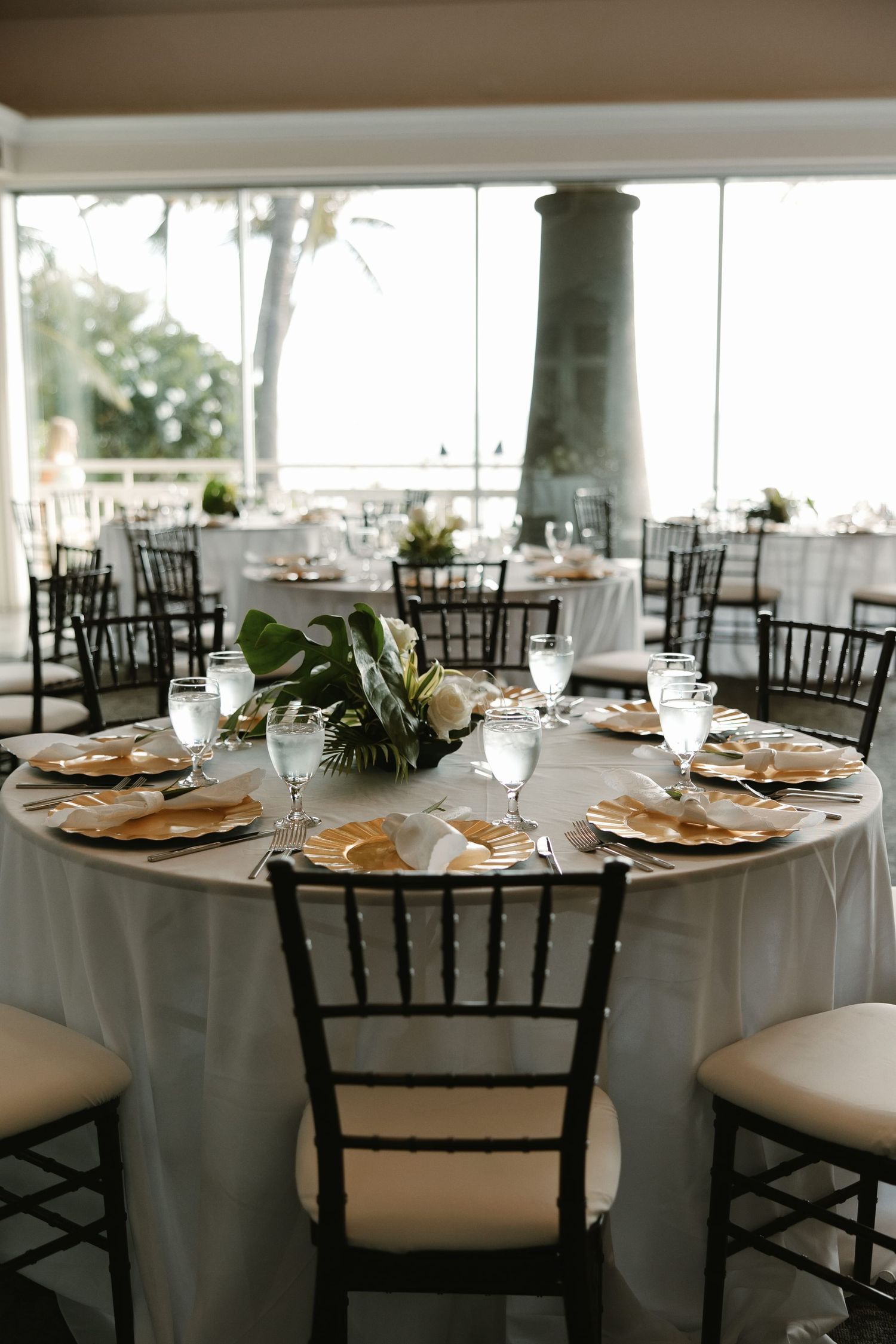 Elegant wedding reception tables with white linens, gold chargers, floral centerpieces, and black Chiavari chairs in a bright venue.